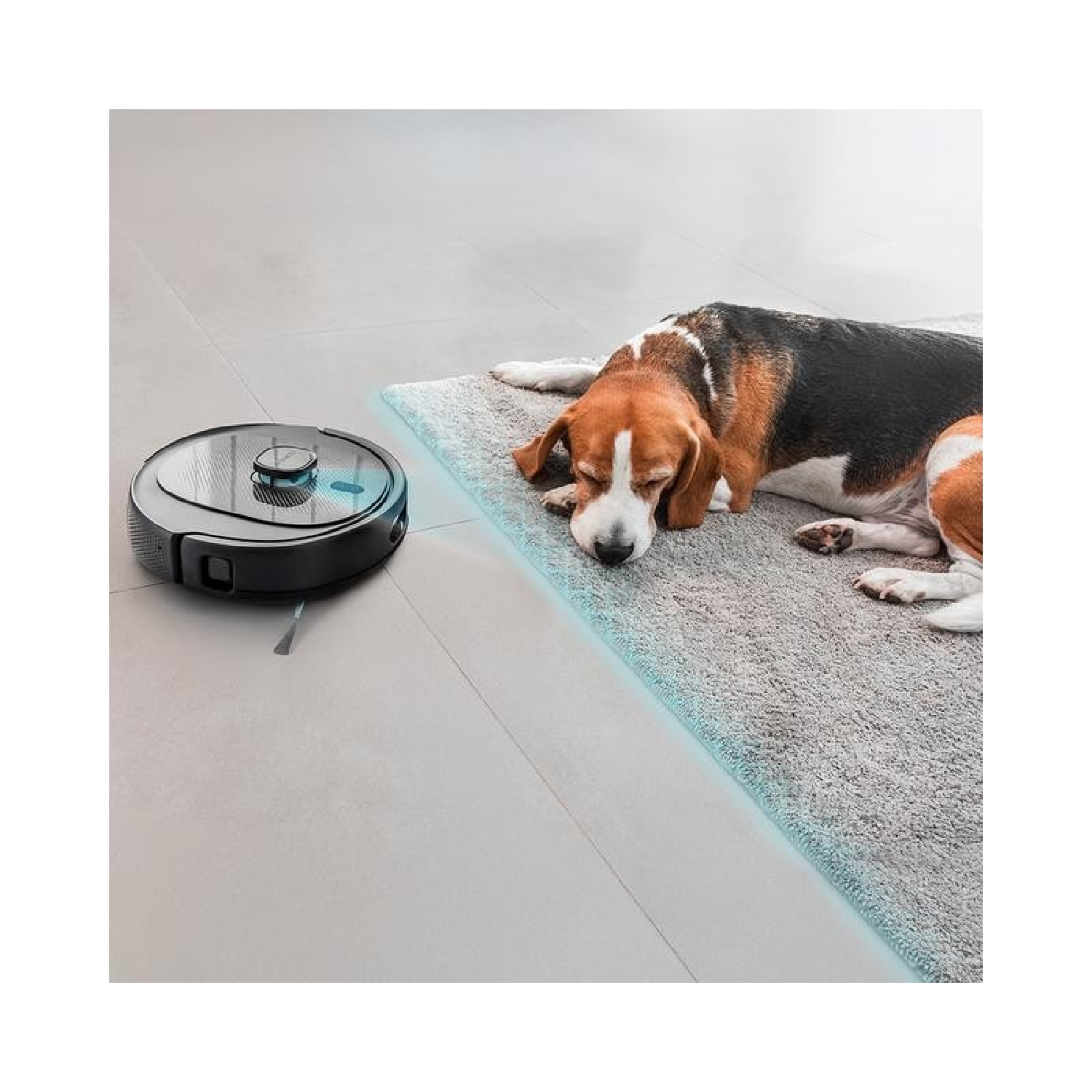 Un robot aspirapolvere accanto a un cane beagle che riposa su un tappeto. Il cane è marrone, nero e bianco.