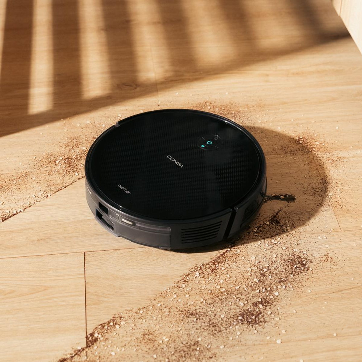 Una aspiradora robot negra en un piso de madera con escombros dispersos. Sombras y una pata de madera al fondo.