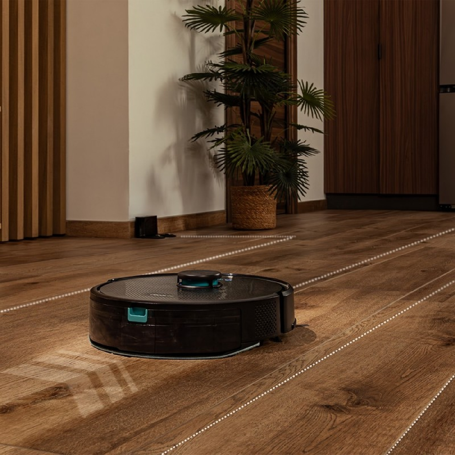 Un robot aspirapolvere su un pavimento in legno. Una pianta in un cesto di vimini sullo sfondo. Si vede una porta in legno scuro.