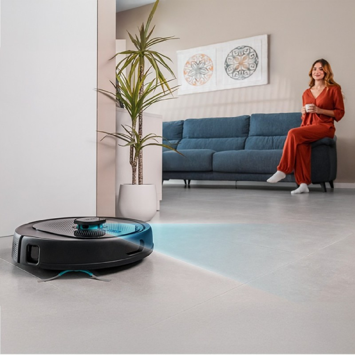 Una mujer sentada en un sofá. Una aspiradora robot limpia el suelo. Una planta en maceta. La habitación tiene un diseño moderno.