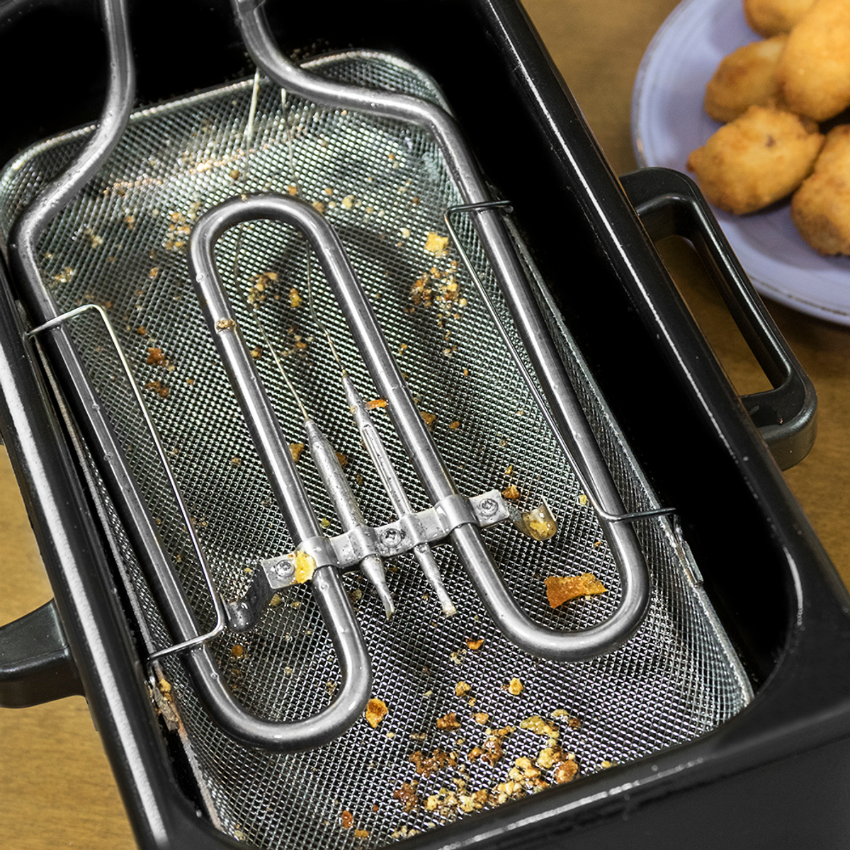 Primer plano de una cesta de freidora con un elemento calefactor. La cesta contiene restos. Nuggets en un plato al fondo.