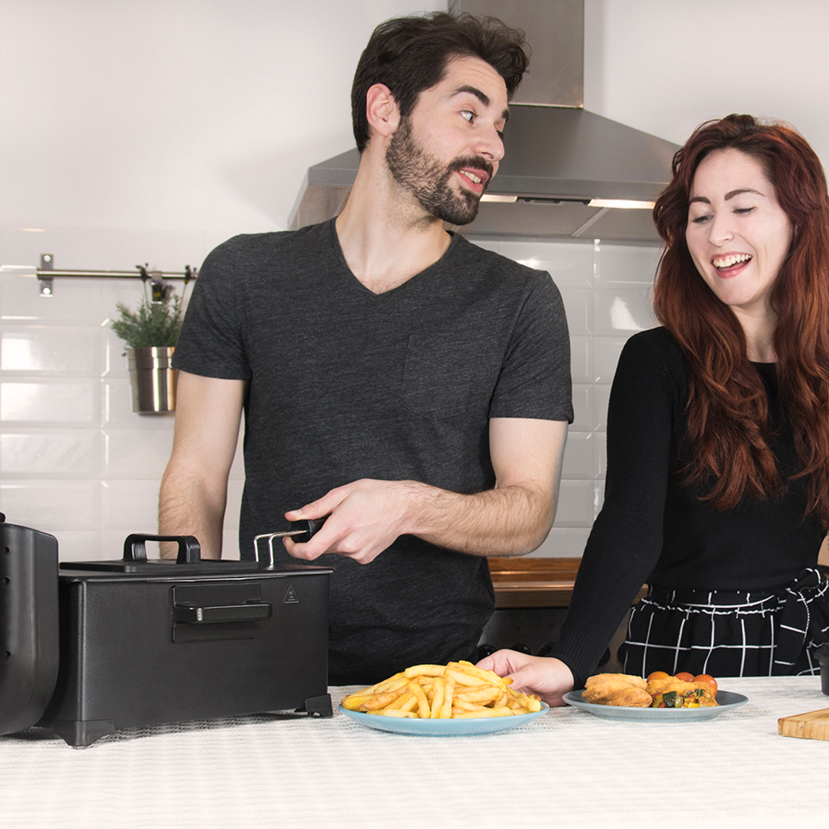 Un hombre y una mujer en una cocina, sonriendo. Una freidora, platos de comida y una encimera están a la vista. La escena sugiere cocinar.