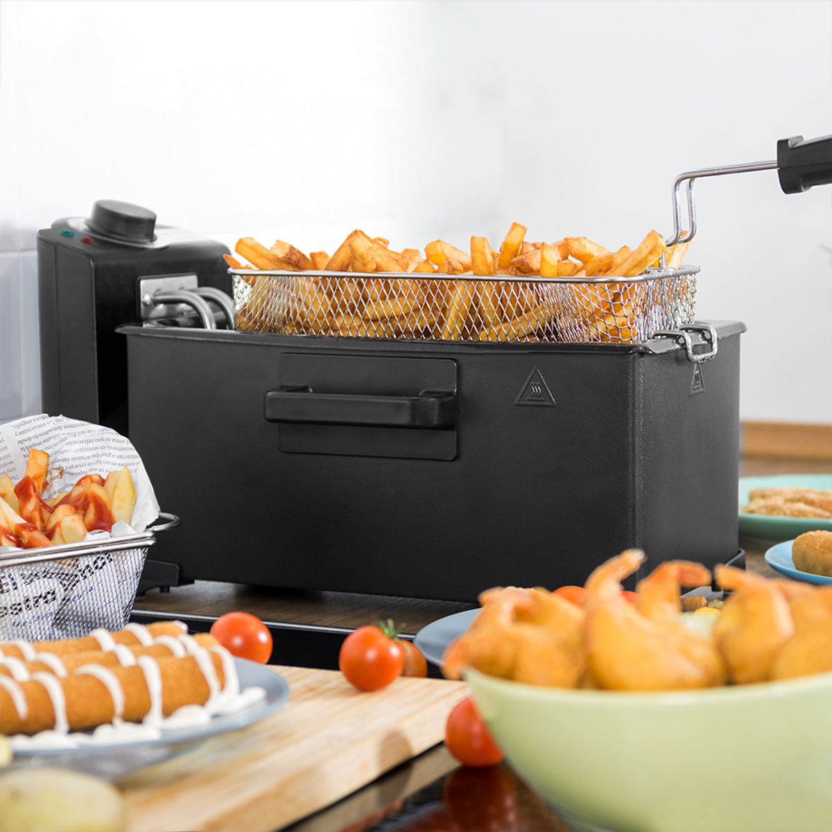 Una freidora con una cesta de patatas fritas está sobre una encimera. Otros platos, tomates y un bol de comida están cerca.