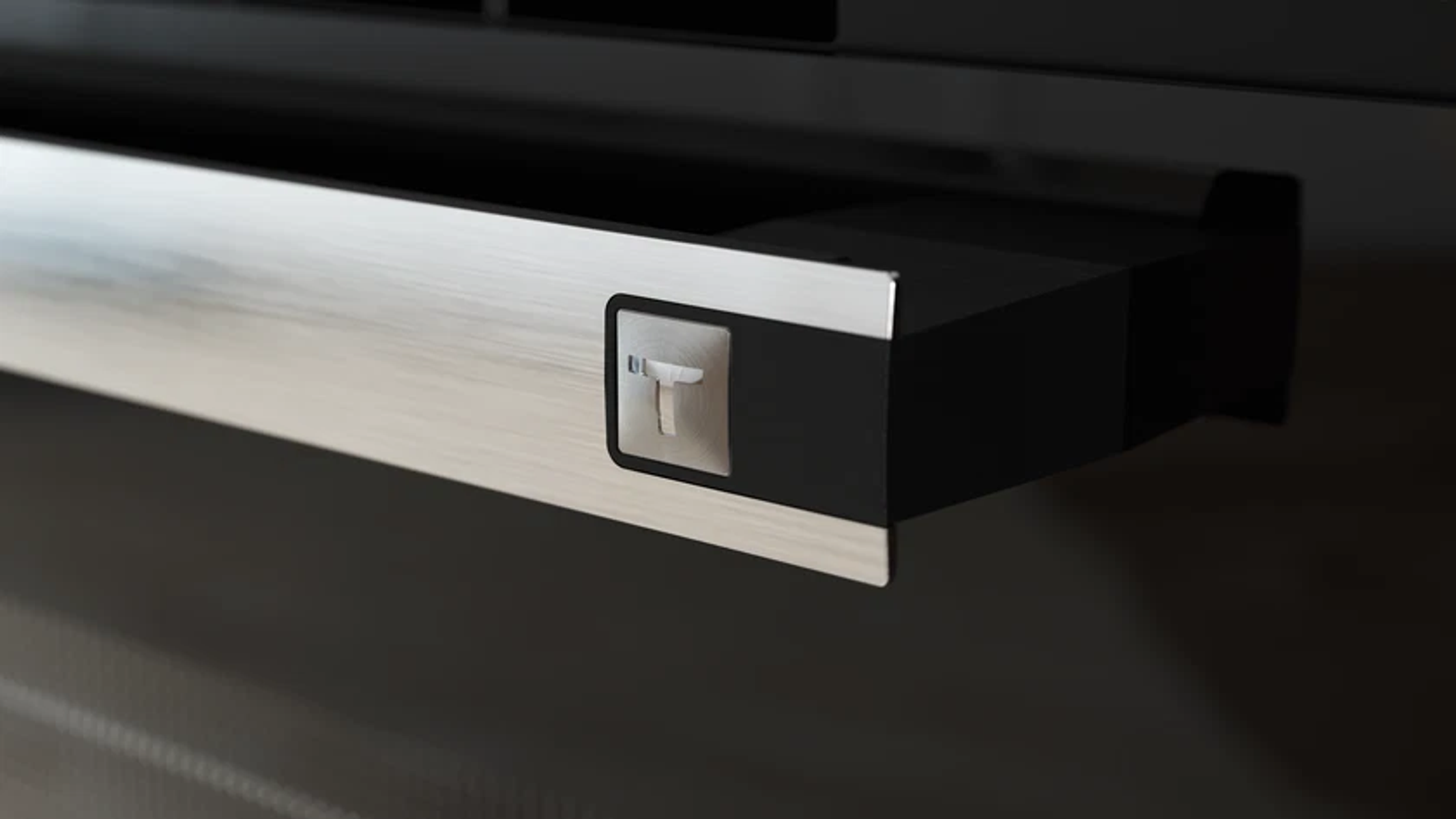 Primer plano de un tirador de cajón de acero inoxidable con borde negro, que muestra un botón en forma de 'T' para abrir.