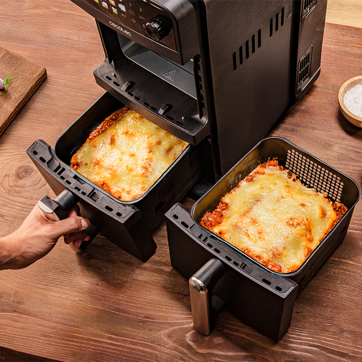 Una friggitrice ad aria con due vassoi di lasagne che vengono estratti. Un tavolo di legno con sale e un tagliere sullo sfondo.