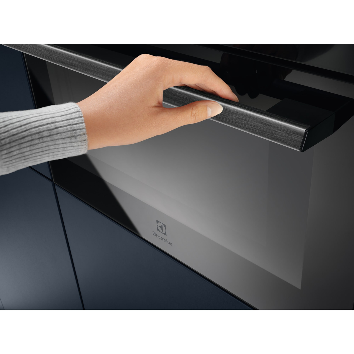 Una mano abre una puerta de horno negra con un asa metálica. El horno tiene una puerta de vidrio y el logotipo de Electrolux es visible.