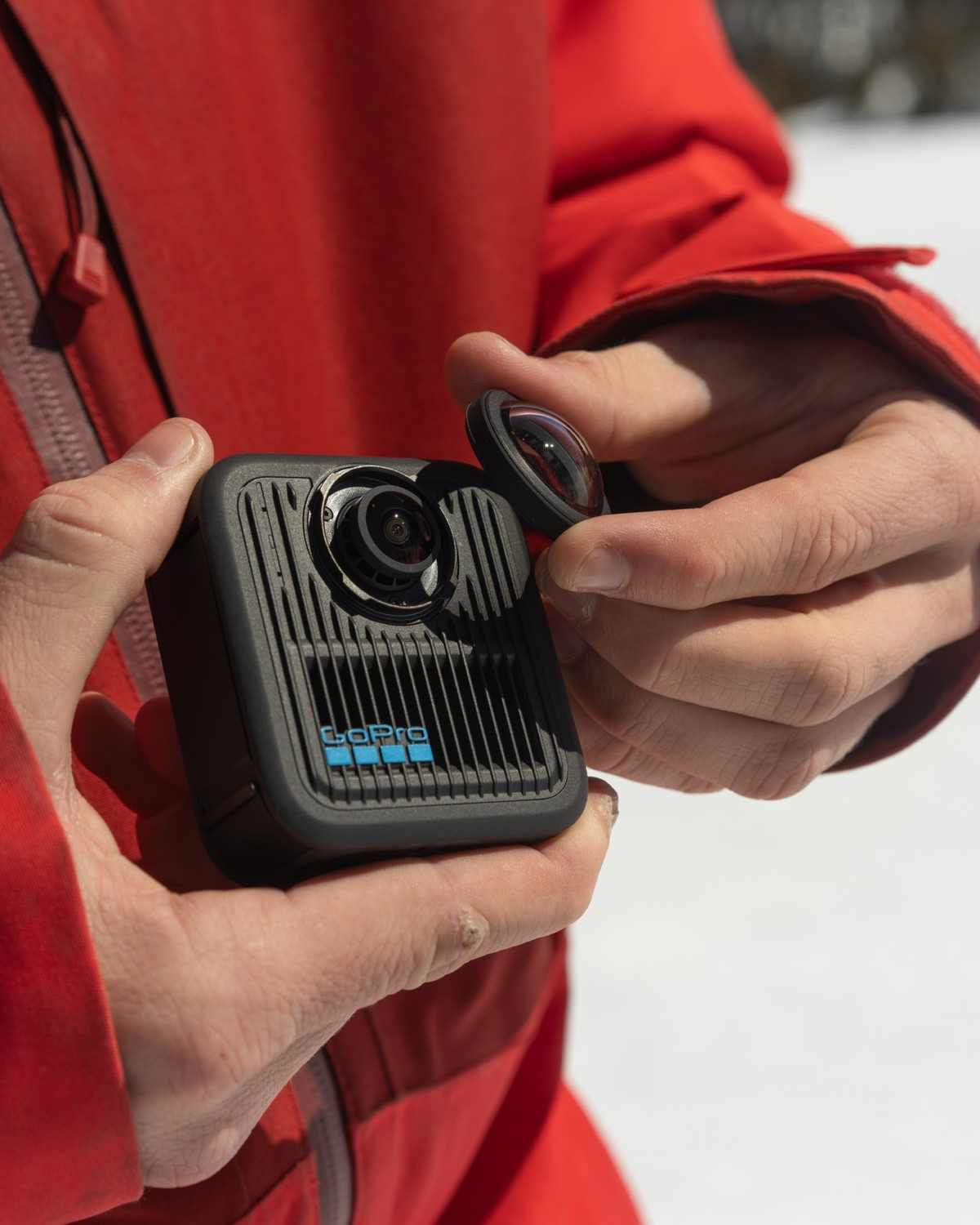 Eine Person hält eine GoPro-Kamera und -Linse. Die Kamera ist schwarz mit blauem GoPro-Logo. Die Person trägt eine rote Jacke, Schnee im Hintergrund.