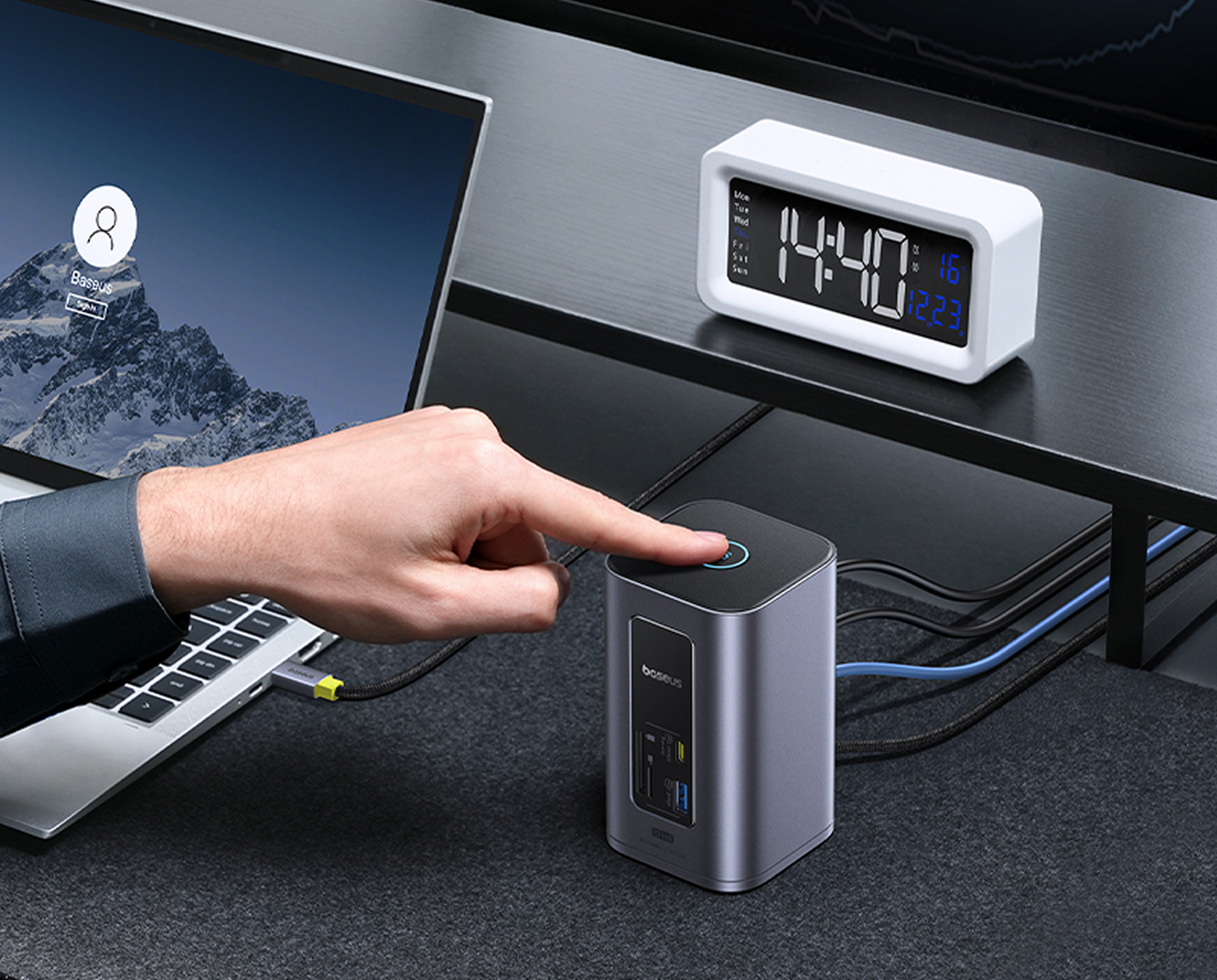 Una mano preme un pulsante su un dispositivo grigio. Un laptop, un orologio bianco e dei cavi sono su una scrivania.