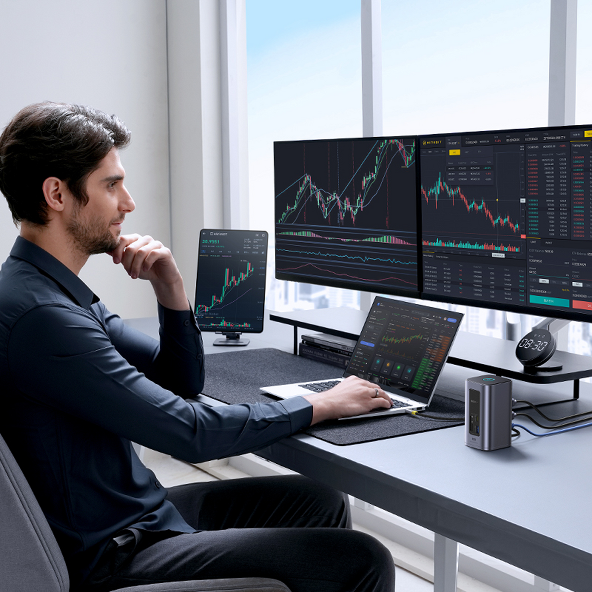 Uomo in camicia scura alla scrivania con schermi che mostrano grafici di borsa, usa laptop e tablet.