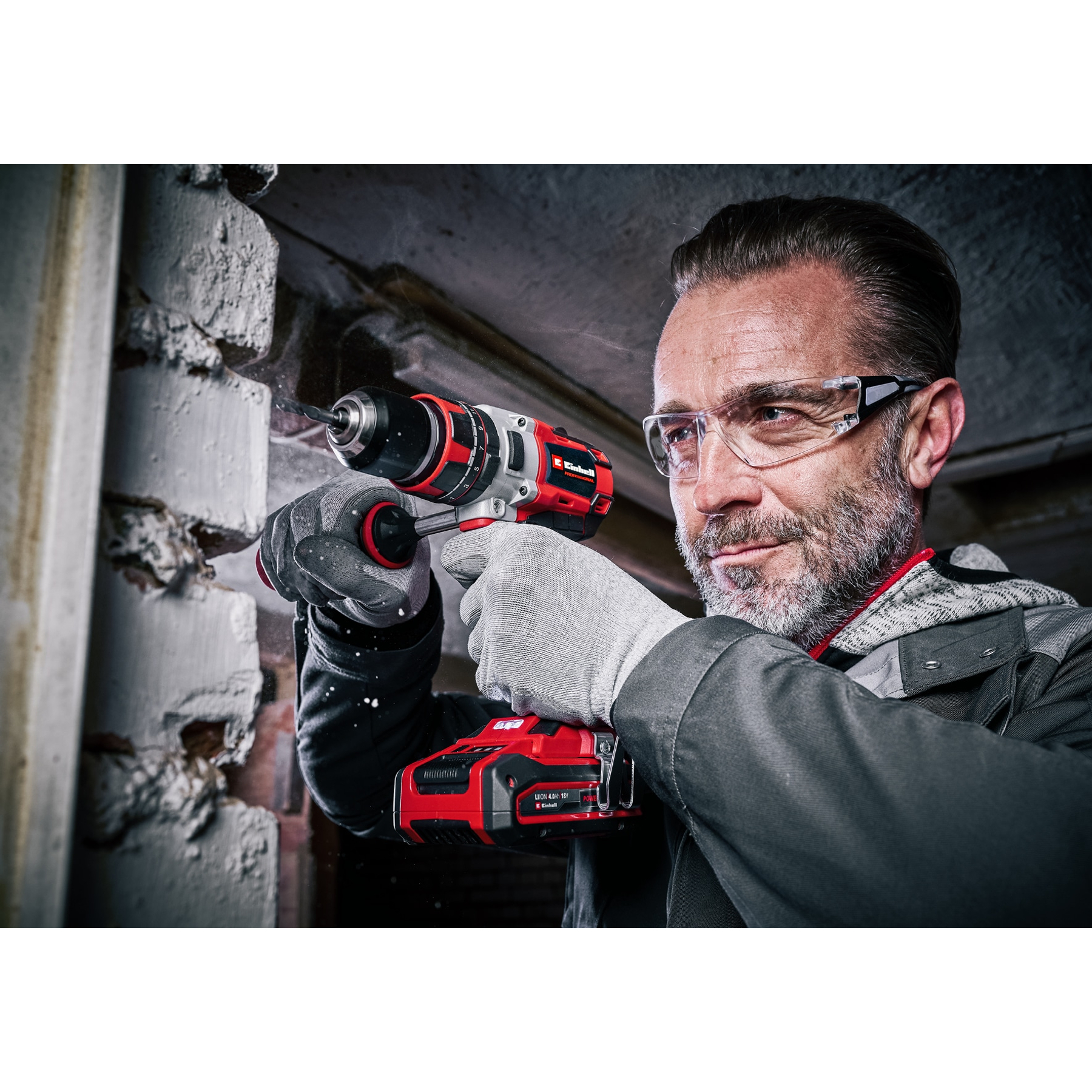 Mężczyzna wierci w ceglanym murze czerwoną i czarną wiertarką, w okularach ochronnych i rękawicach.