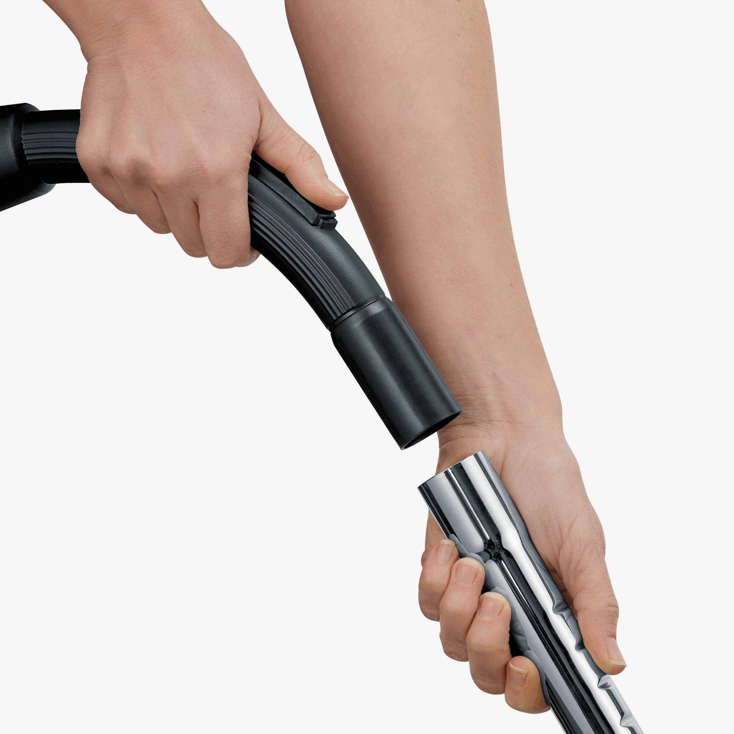 Ręce łączą czarny wąż odkurzacza z błyszczącą srebrną rurą przedłużającą. Białe tło.