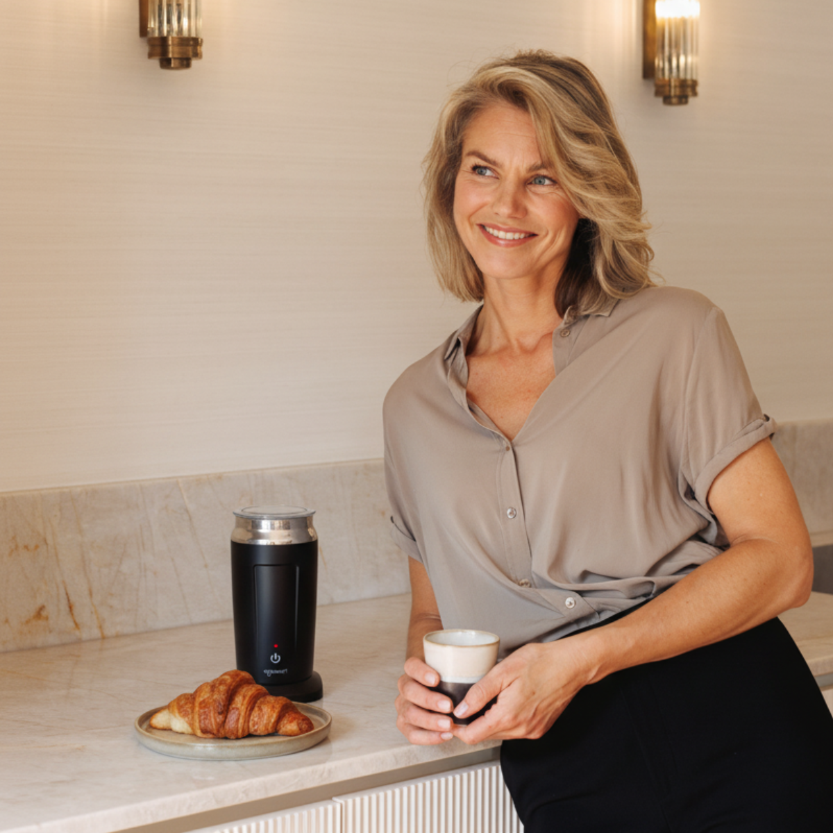 Een vrouw glimlacht, met een kopje in haar hand. Een croissant en een koffiezetapparaat staan op het aanrecht. Beige shirt en zwarte broek.