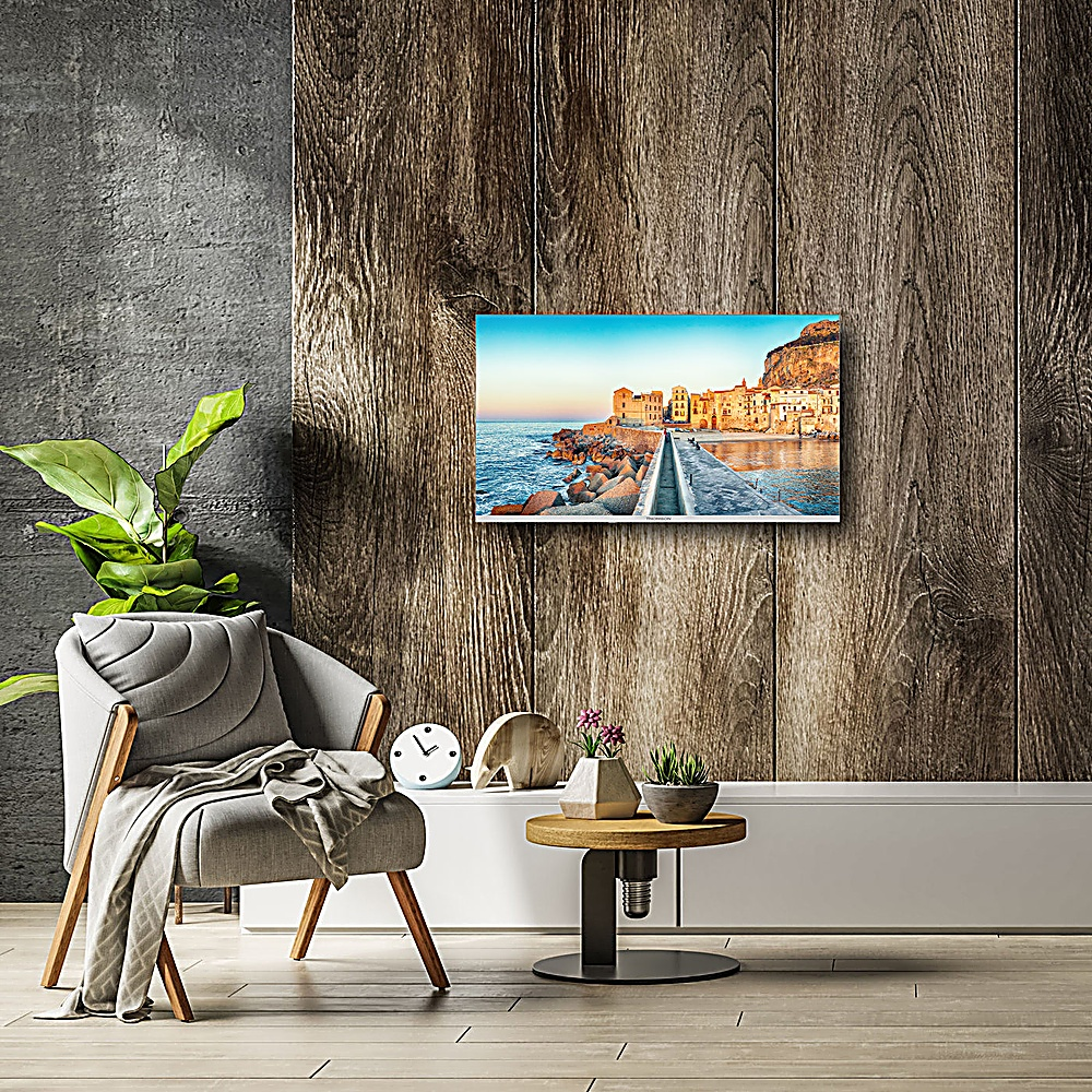 Un'immagine incorniciata di una città costiera è appesa a una parete rivestita di legno. Una sedia grigia e un tavolino con decorazioni sono in primo piano.