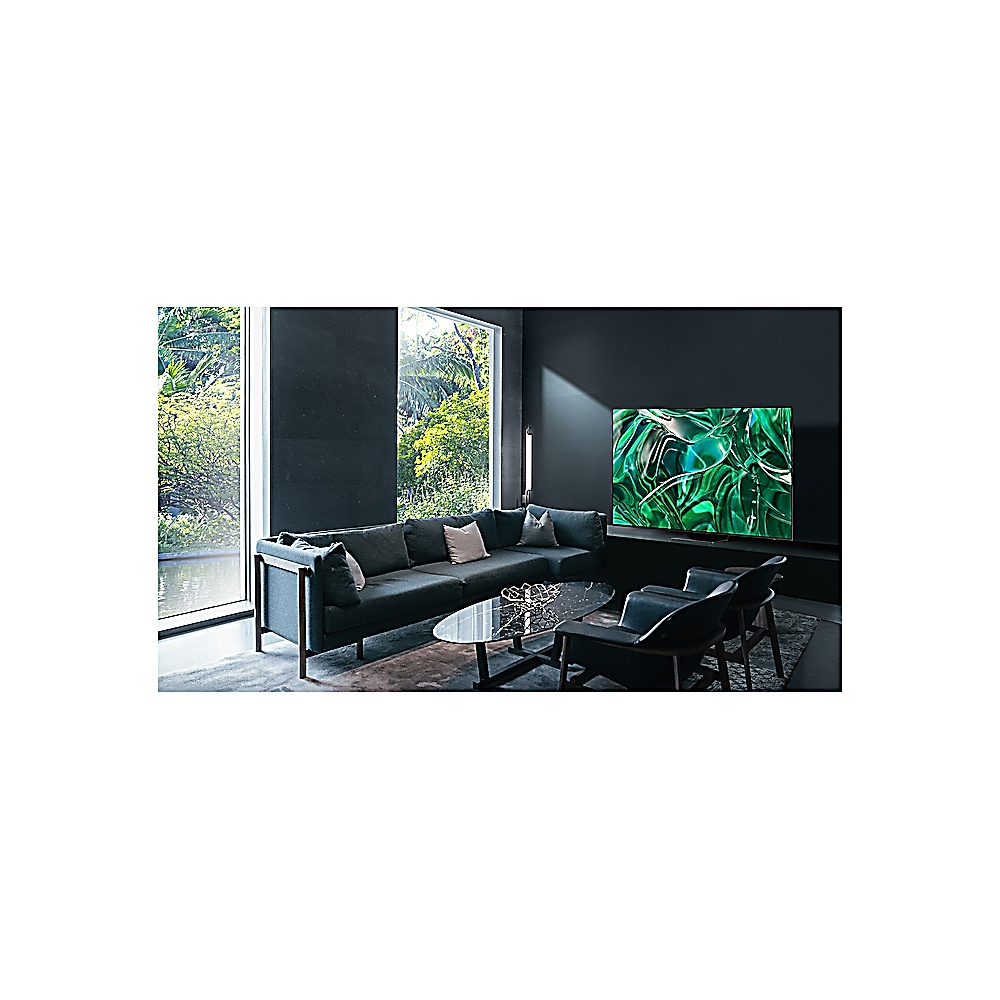 Salon avec canapé gris foncé, table en verre, deux chaises et une grande télévision. Une fenêtre montre un paysage vert.