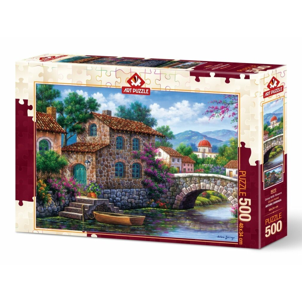 Una scatola di puzzle che mostra una scena di villaggio con una casa in pietra, un ponte e una barca. Il cielo è blu con nuvole.