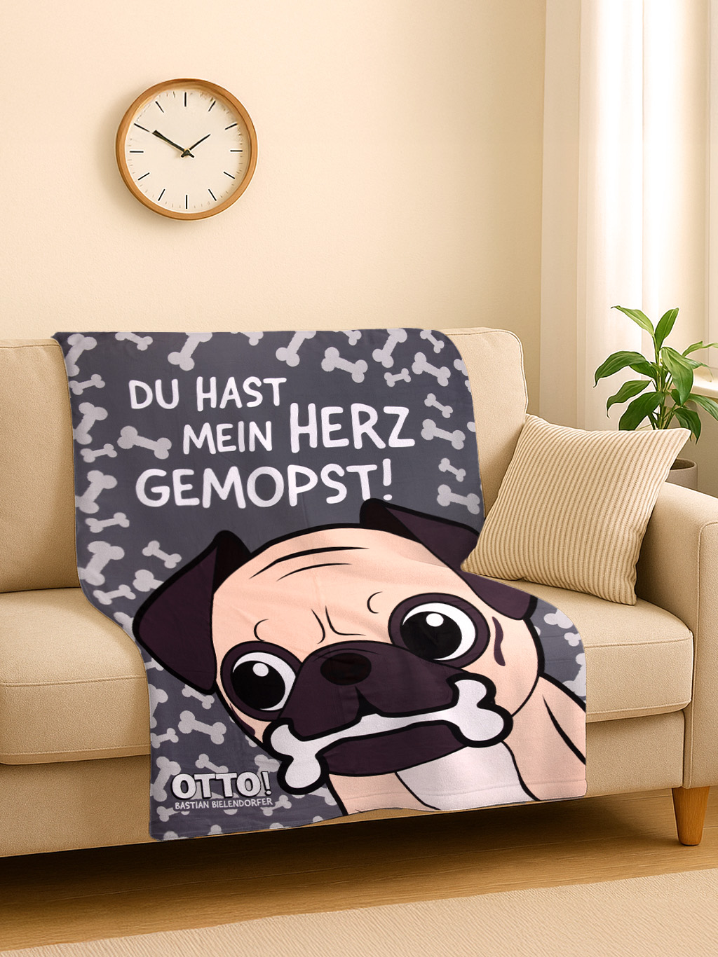 Eine Decke mit einer Mops-Illustration liegt auf einem Sofa. Die Decke hat Text, Knochenmuster und eine Uhr hängt an der Wand.