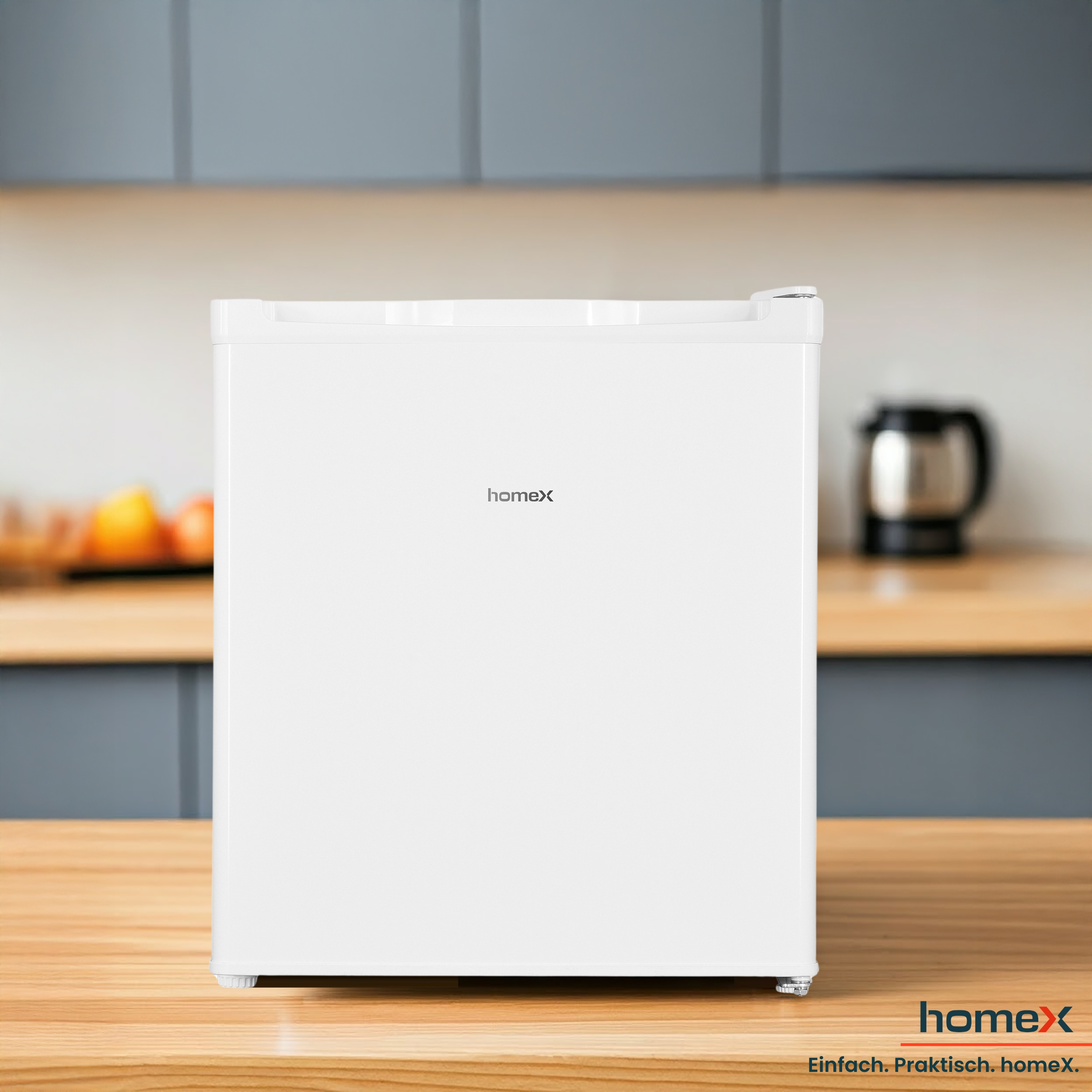 Ein weißer Mini-Kühlschrank mit dem Wort 'homex' steht auf einer Holztheke. Ein schwarzer Wasserkocher ist im Hintergrund.