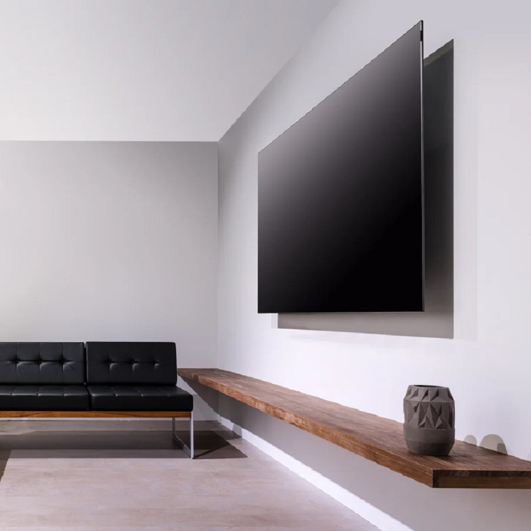 Salón moderno con TV grande en pared blanca. Se ven estante de madera y sofá de cuero negro.