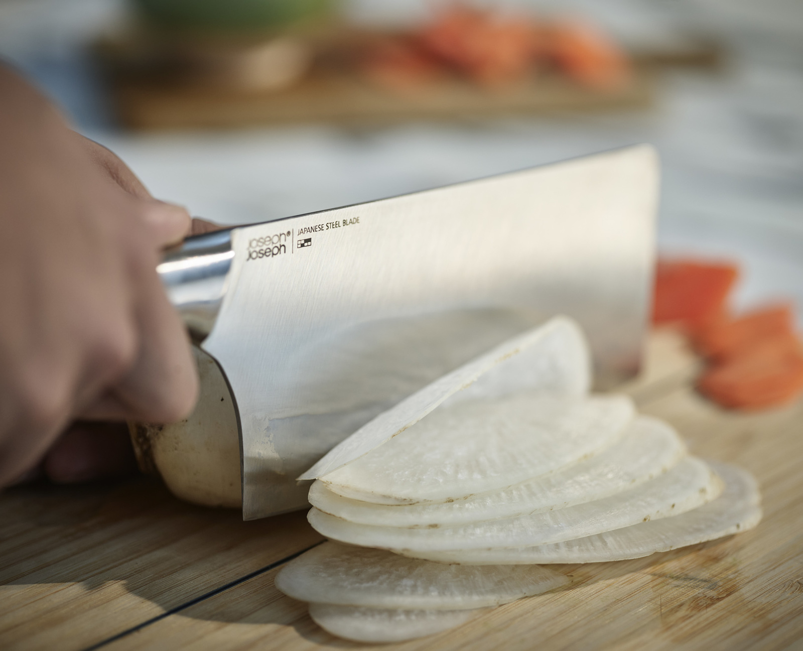 Una mano usa un cuchillo de acero japonés para cortar un rábano blanco sobre una tabla de cortar de madera. Zanahorias cortadas en el fondo.