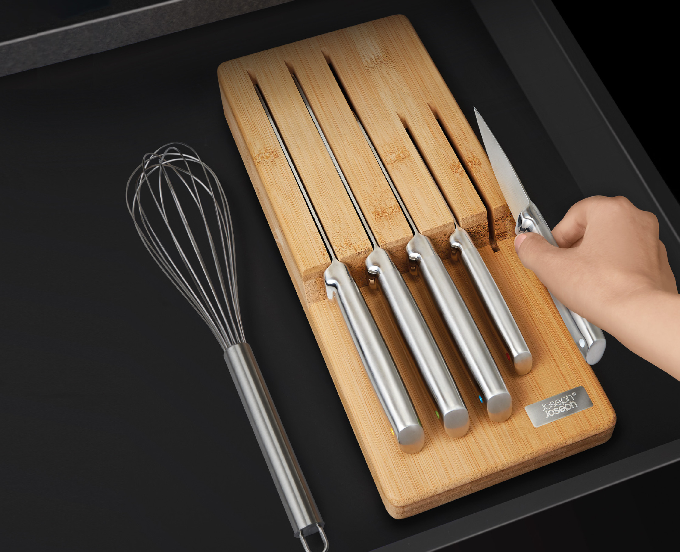 Un cajón de cocina contiene cuchillos en un bloque de bambú, un batidor y una mano que alcanza un cuchillo. Fondo negro.