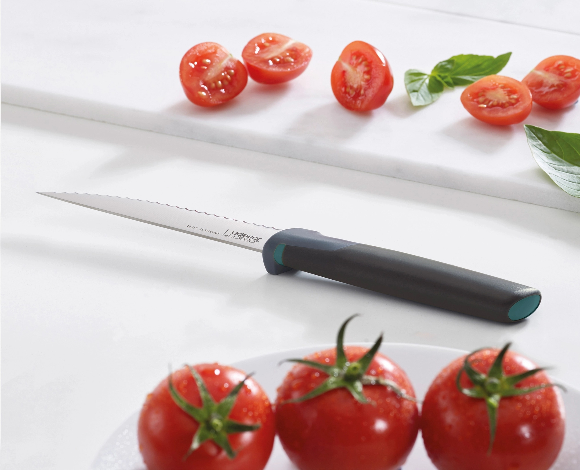 Tomates, cuchillo y albahaca sobre una superficie blanca. El cuchillo está en el centro con tomates arriba y abajo.