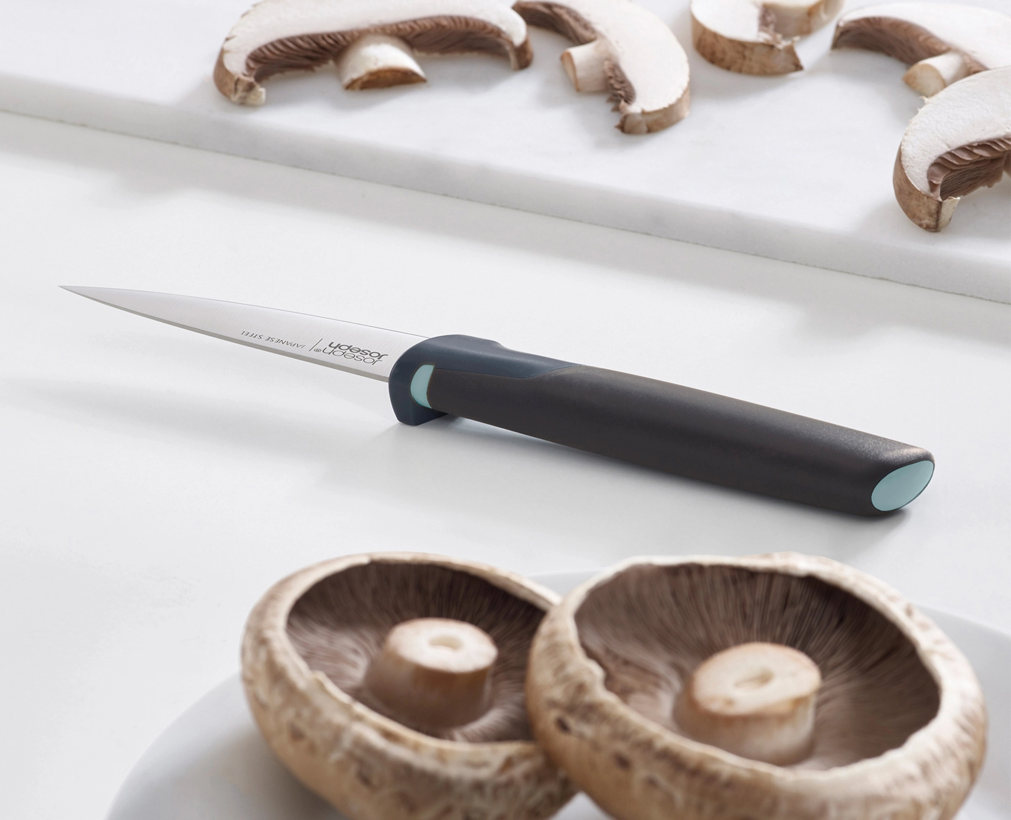 Un cuchillo y champiñones en rodajas sobre una superficie blanca. Dos champiñones enteros en primer plano.