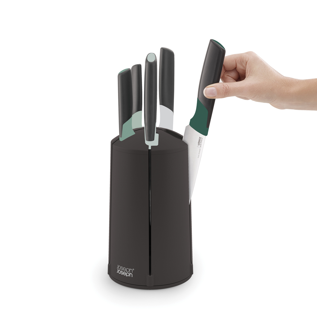 Una mano saca un cuchillo con mango verde de un bloque de cuchillos negro que contiene otros cuchillos. Fondo blanco.