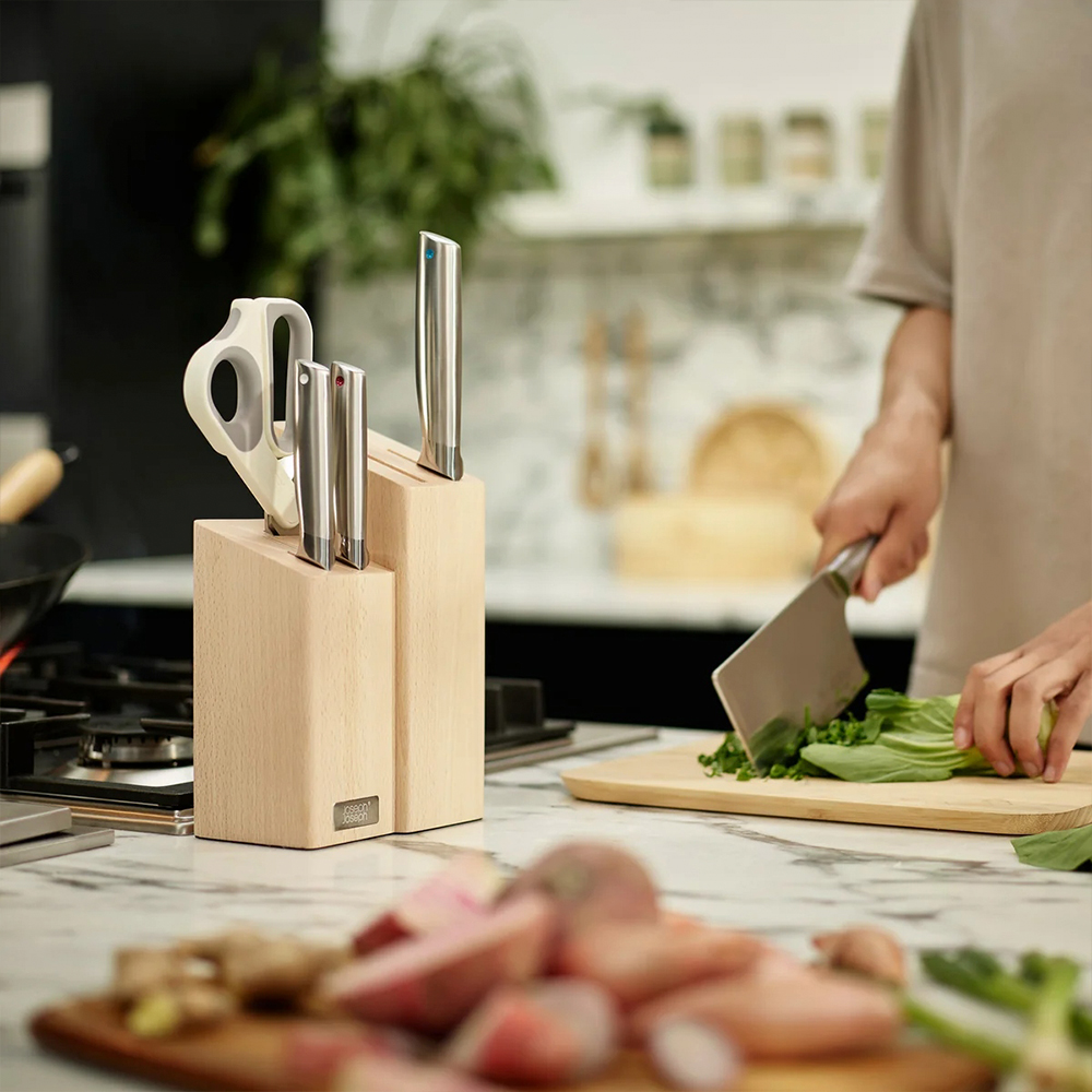 Una persona corta verduras en una tabla de madera con un cuchillo grande. Cuchillos en un bloque de madera están en el mostrador.