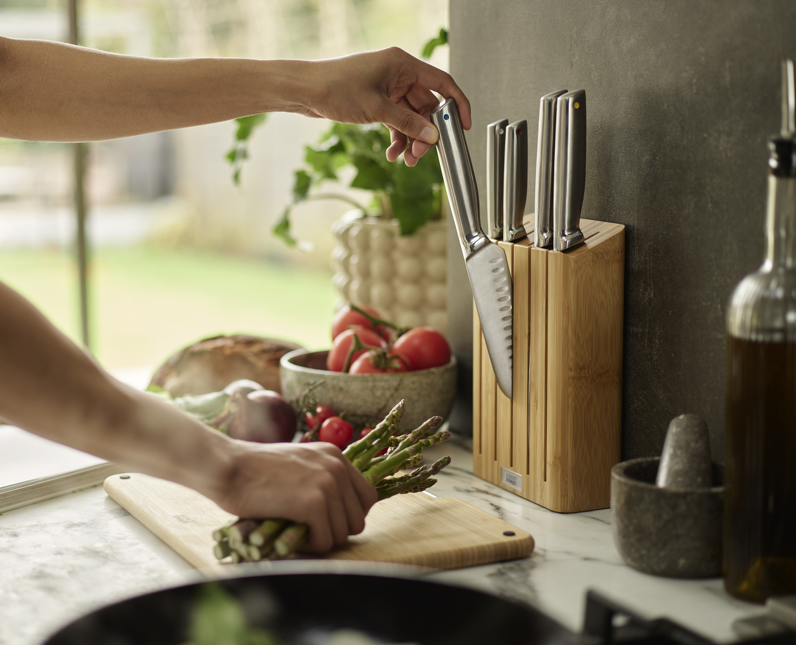 Una persona prepara comida con espárragos y cuchillos. Tomates y pan en el fondo. Se está guardando un cuchillo.