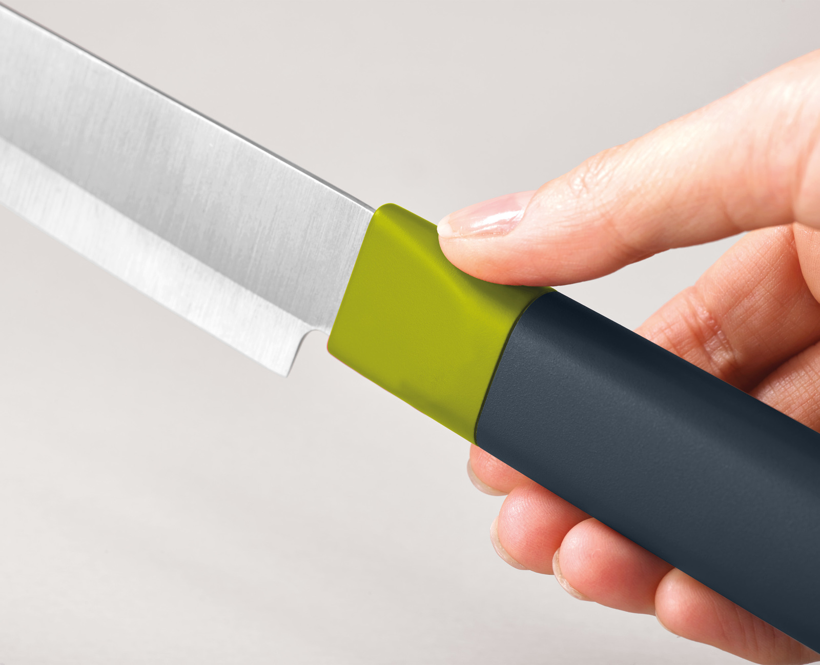 Una mano sostiene un cuchillo. La hoja es plateada. El mango es gris oscuro con un acento verde. El fondo está borroso.