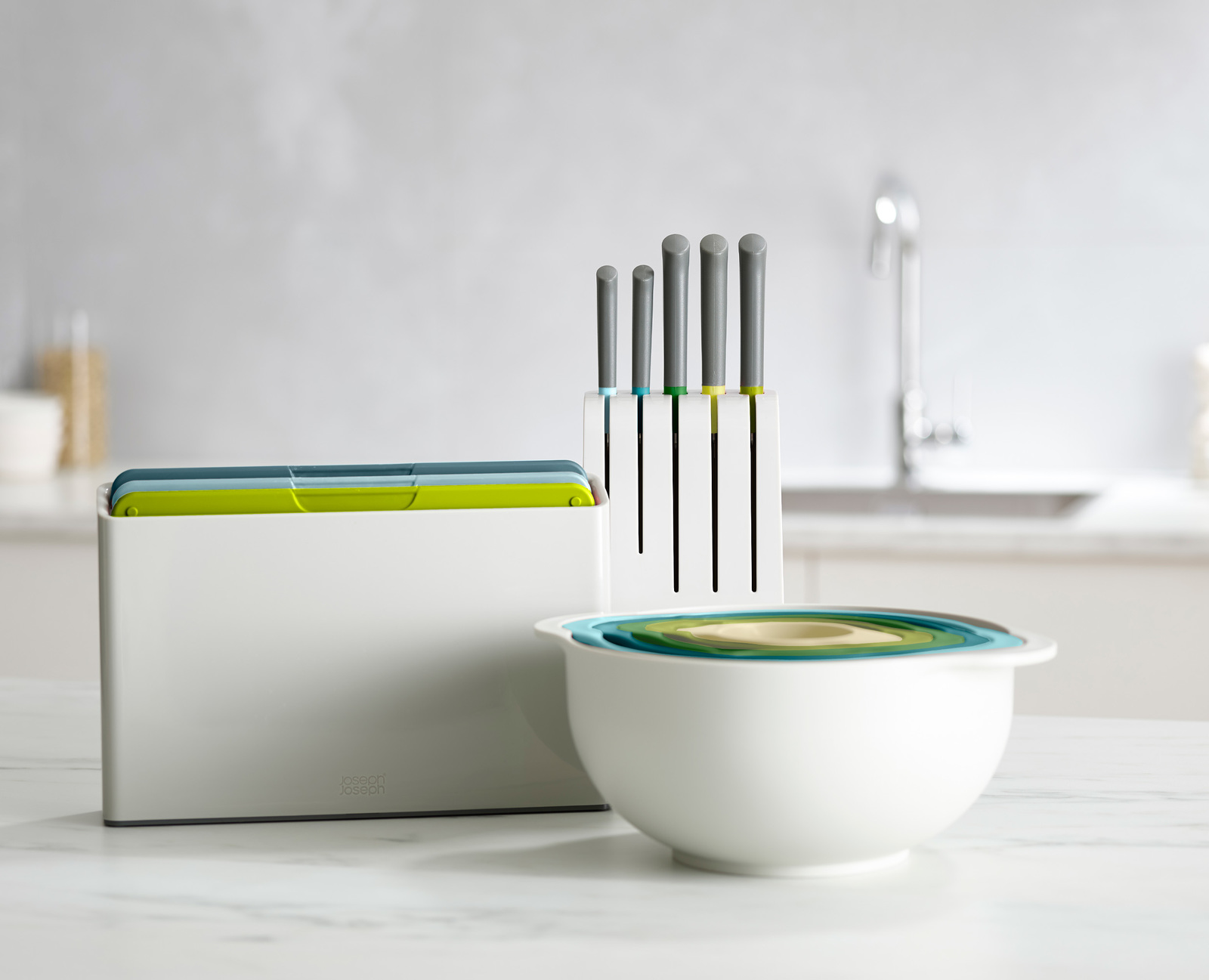 Un juego de cocina blanco con un cuenco, tablas de cortar y cuchillos. El conjunto tiene elementos azules, verdes y blancos. El fondo está borroso.