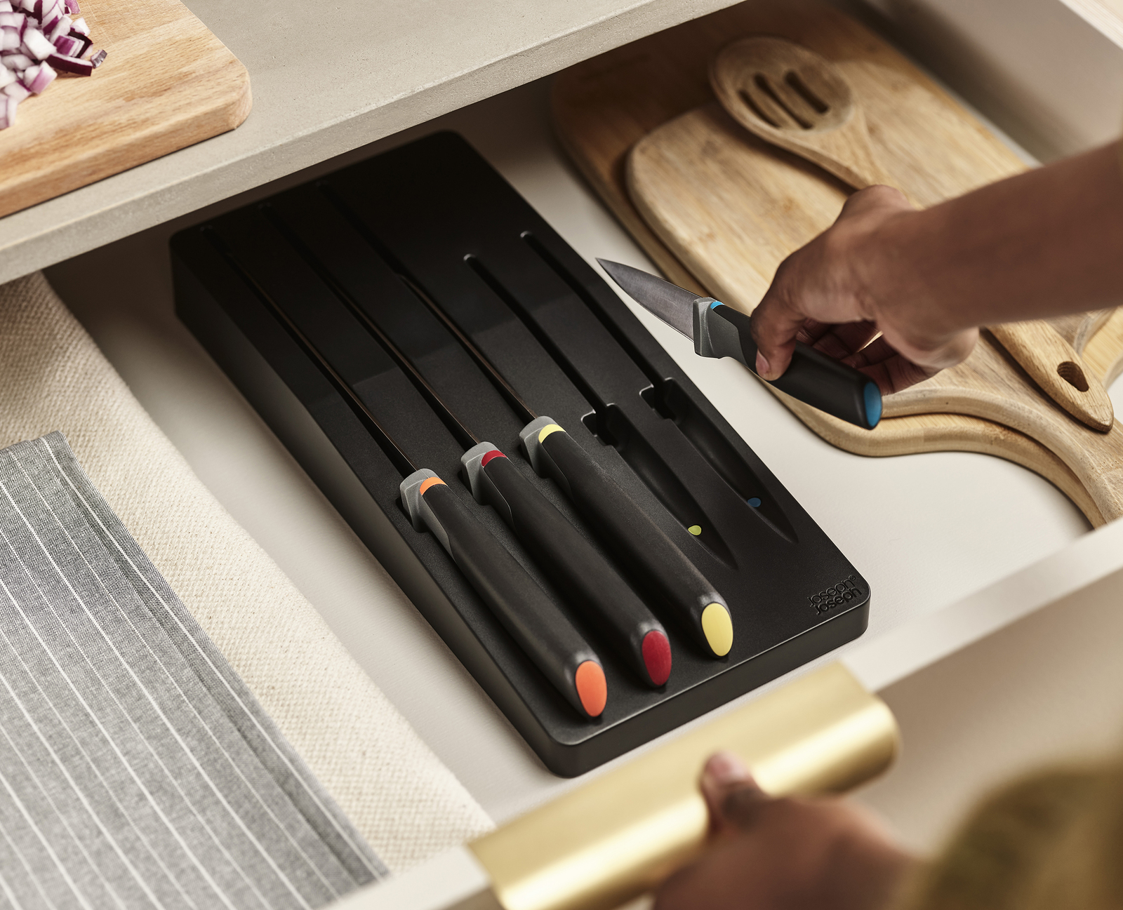 Un cajón con cuchillos en un soporte negro, tablas de cortar y una mano que alcanza un cuchillo. Cebollas picadas en una tabla.