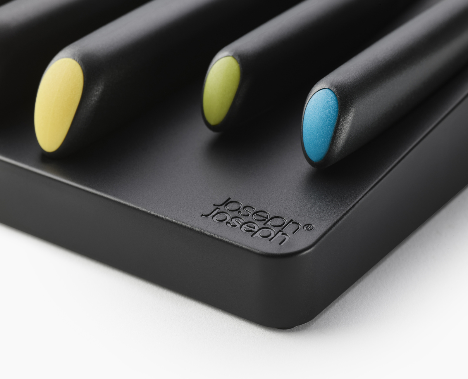 Primer plano de tres cuchillos negros con mangos amarillos, verdes y azules, colocados en un soporte negro con el logo de Joseph Joseph.