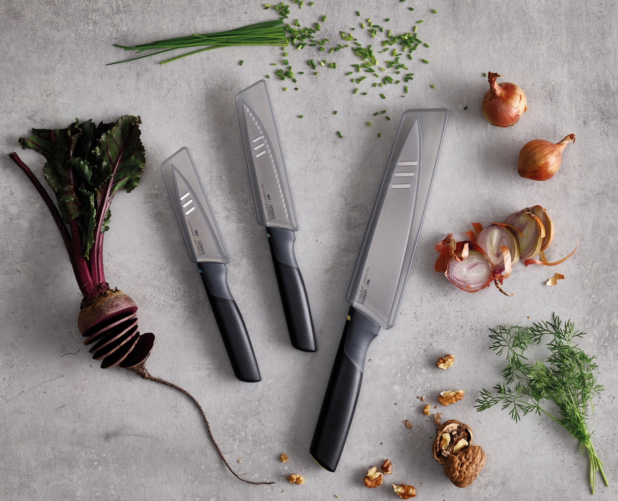 Tres cuchillos con fundas, verduras y nueces sobre una superficie gris, también hay cebollino, remolacha, cebollas y eneldo.