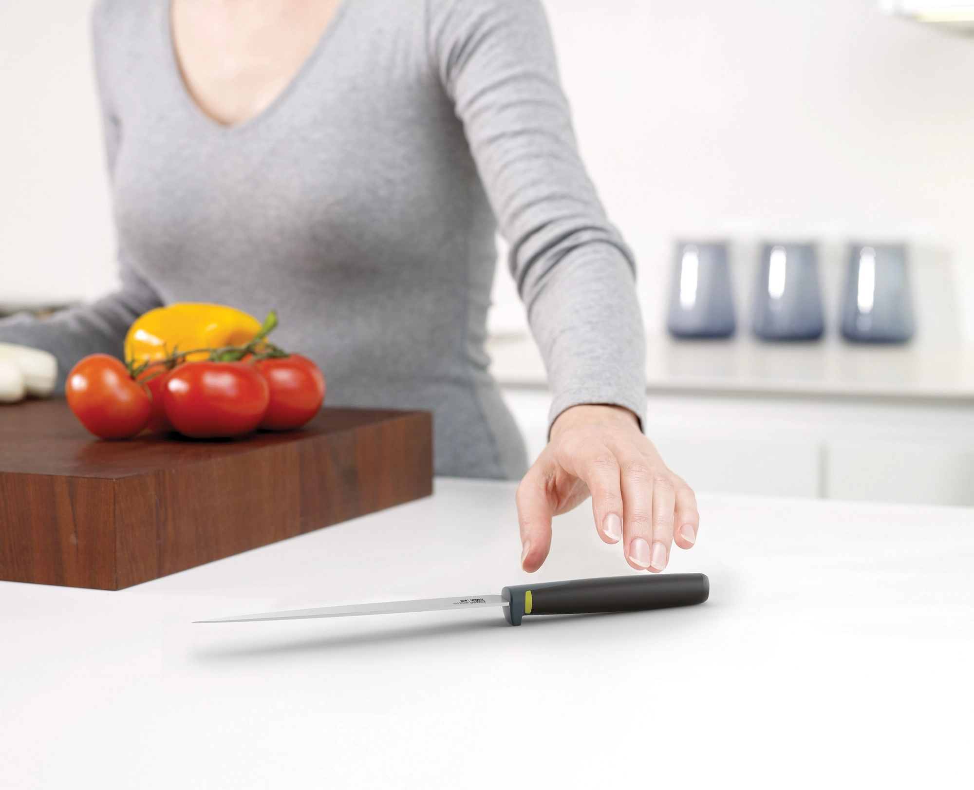 Una persona con una camisa gris alcanza un cuchillo sobre una superficie blanca. Tomates y un pimiento amarillo descansan.