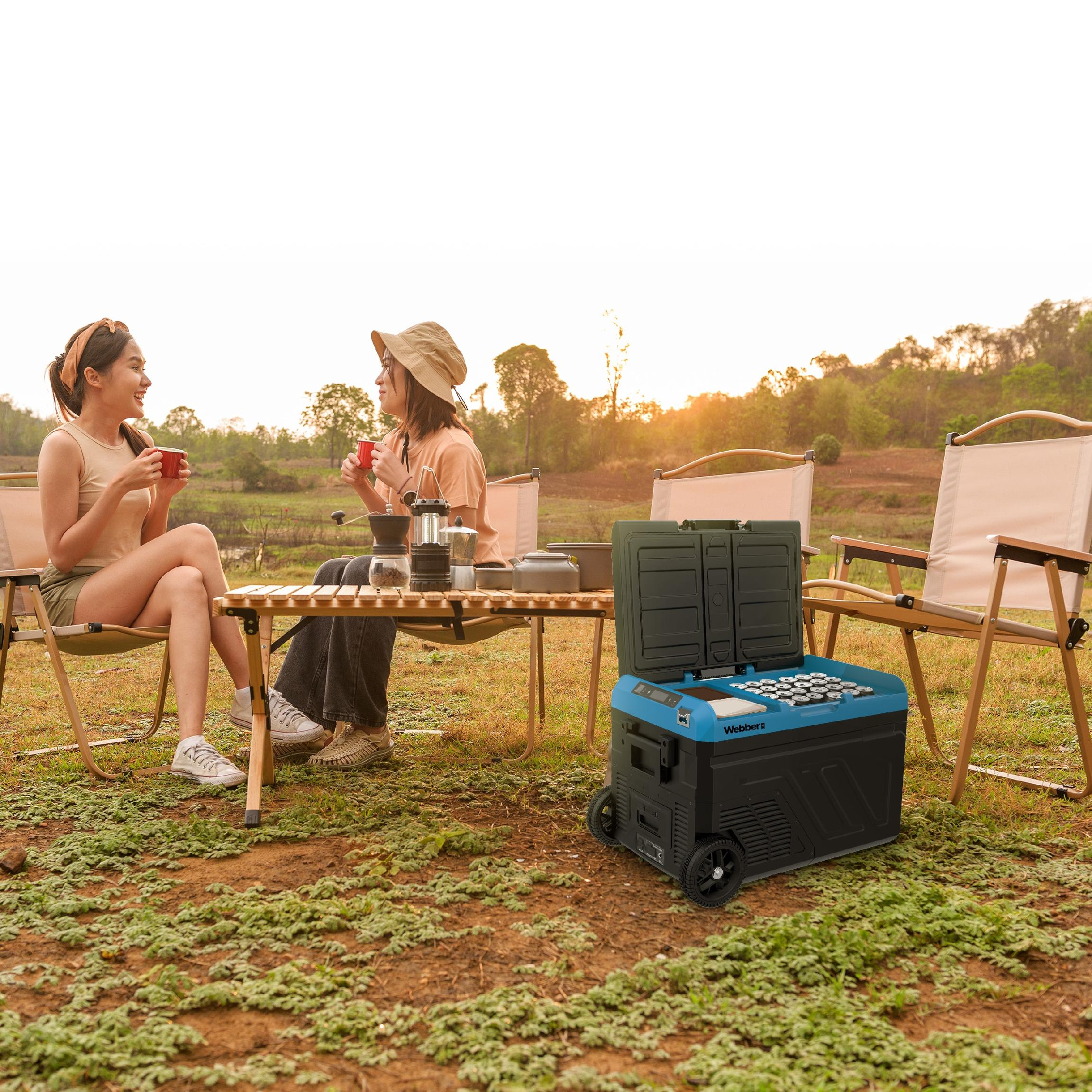 Dwie kobiety na kempingu, siedzące przy stole z kawą. Chłodziarka po prawej. Drzewa i jezioro w tle.
