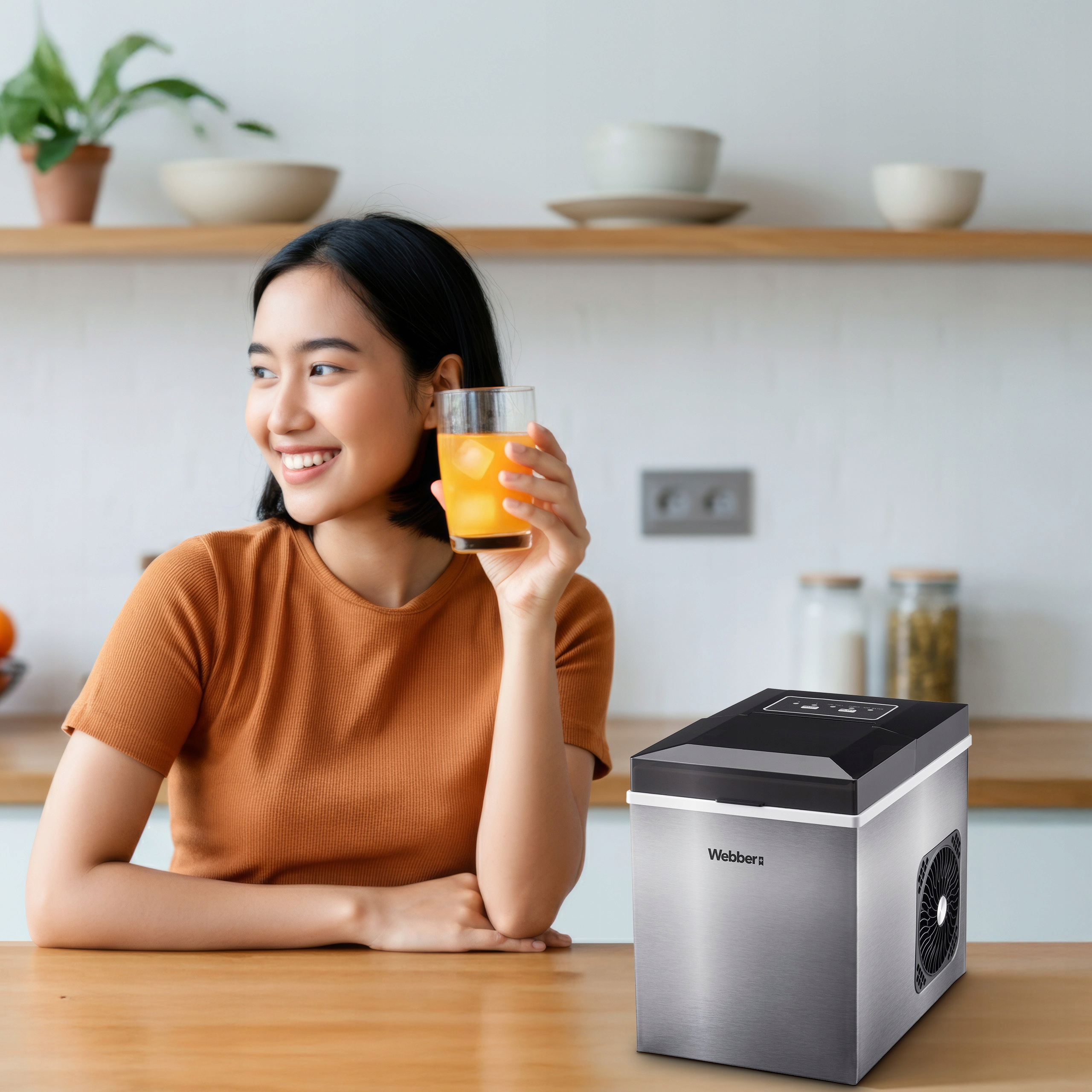 Kobieta uśmiecha się, trzymając szklankę soku pomarańczowego. Wytwornica lodu stoi na stole. Tło kuchni.