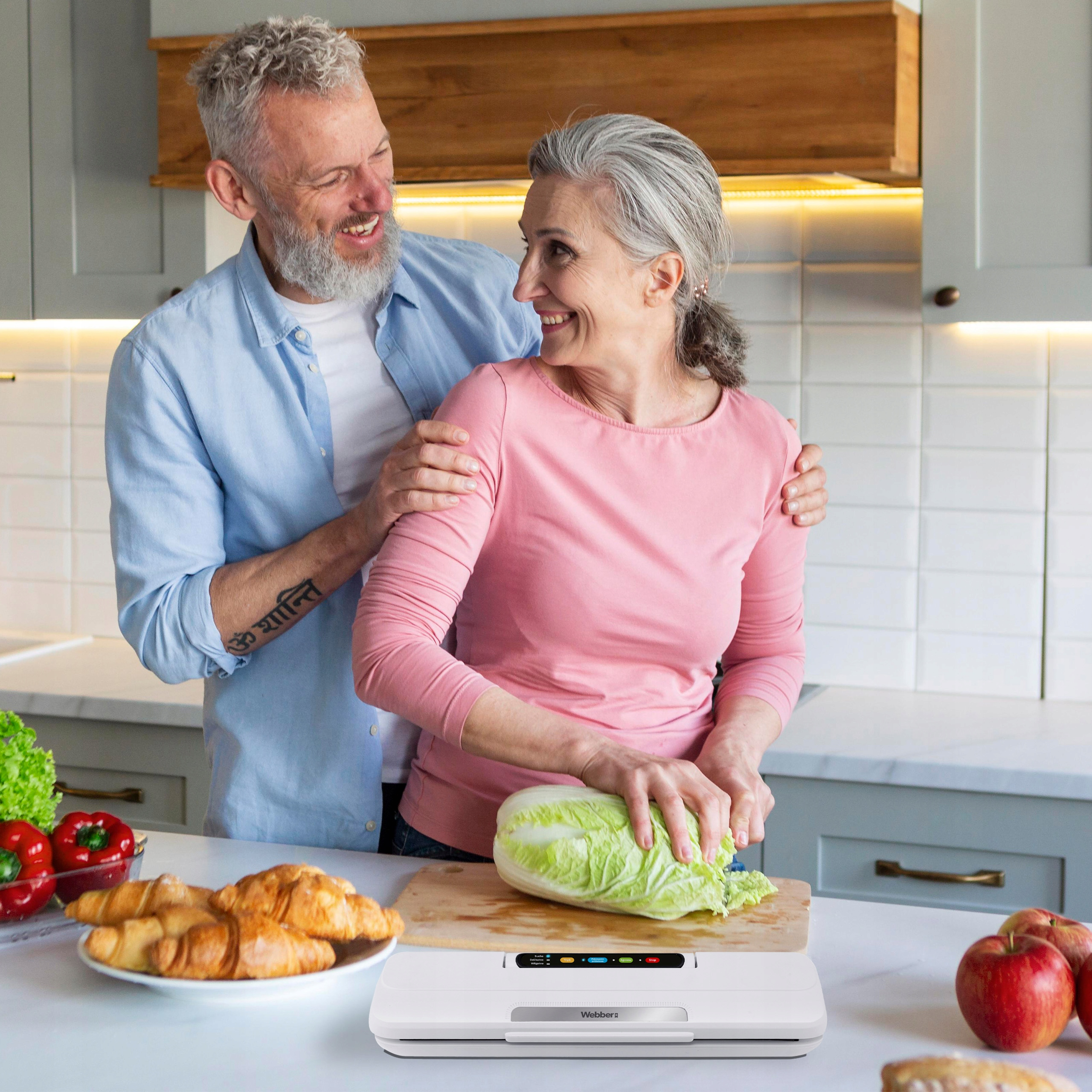 Para w kuchni. Kobieta kroi sałatę; mężczyzna się uśmiecha. Rogaliki, papryka, jabłka i zgrzewarka na blacie.