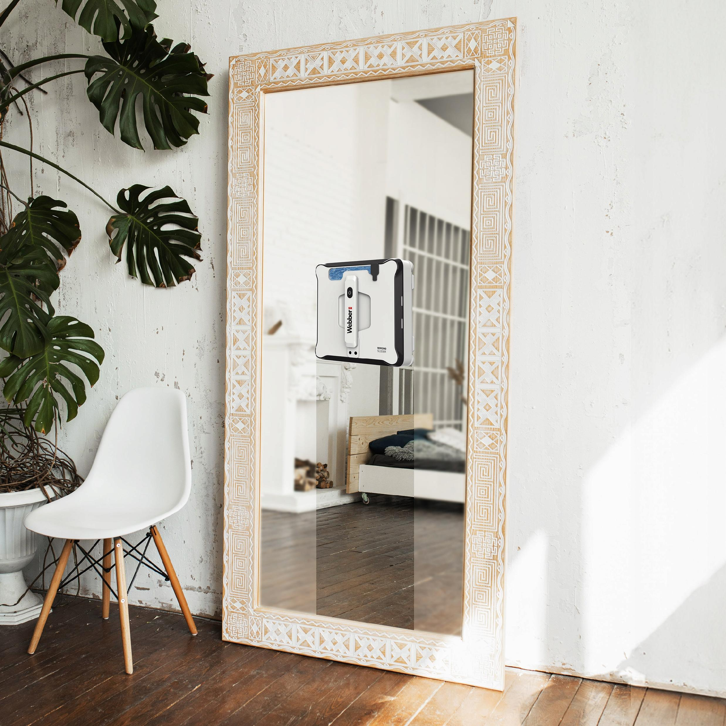 Duże lustro z robotem sprzątającym, białym krzesłem, rośliną i łóżkiem odbitym w lustrze.