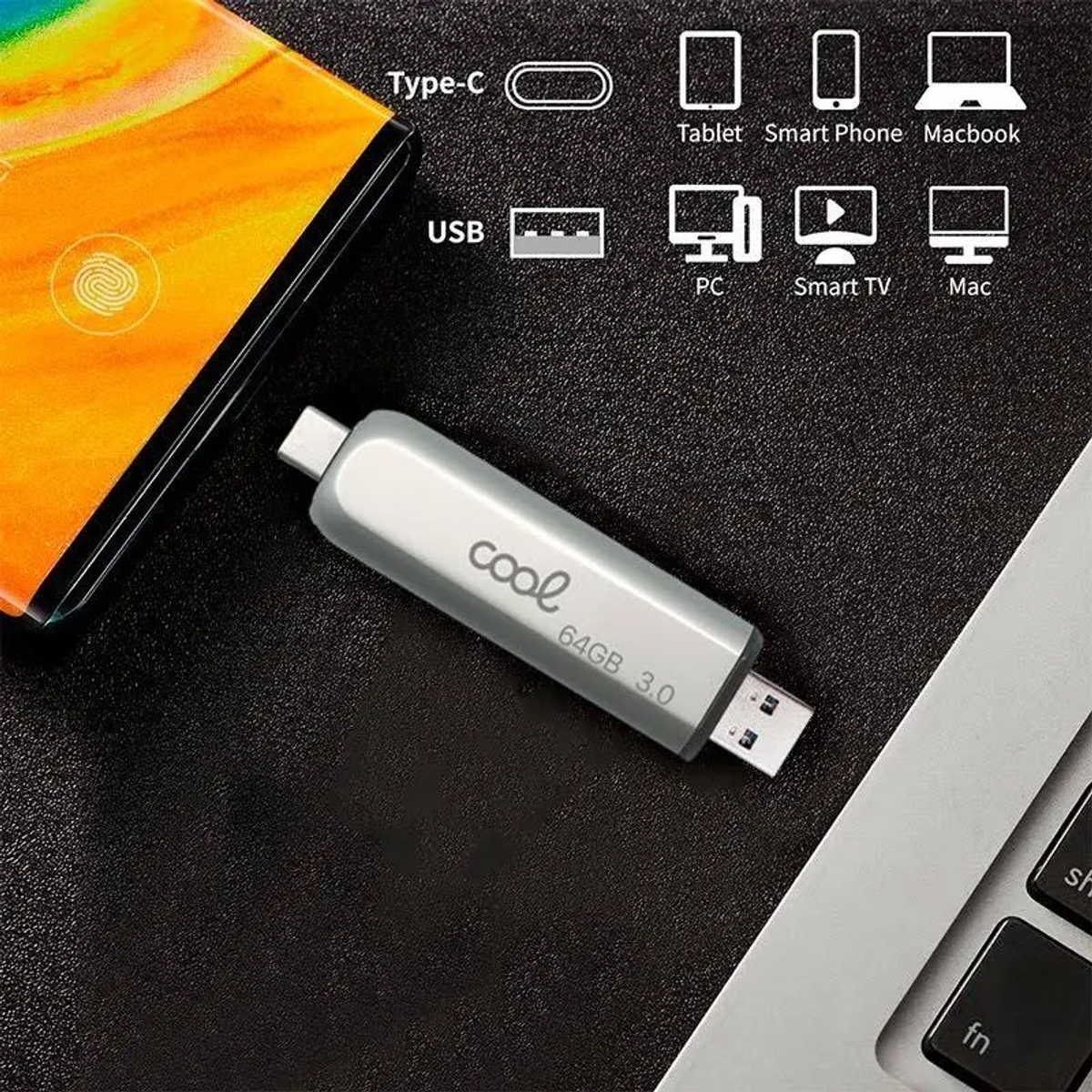 Una unidad USB plateada con 'cool', 64GB 3.0, yace sobre una superficie oscura, cerca de un teléfono y una laptop.