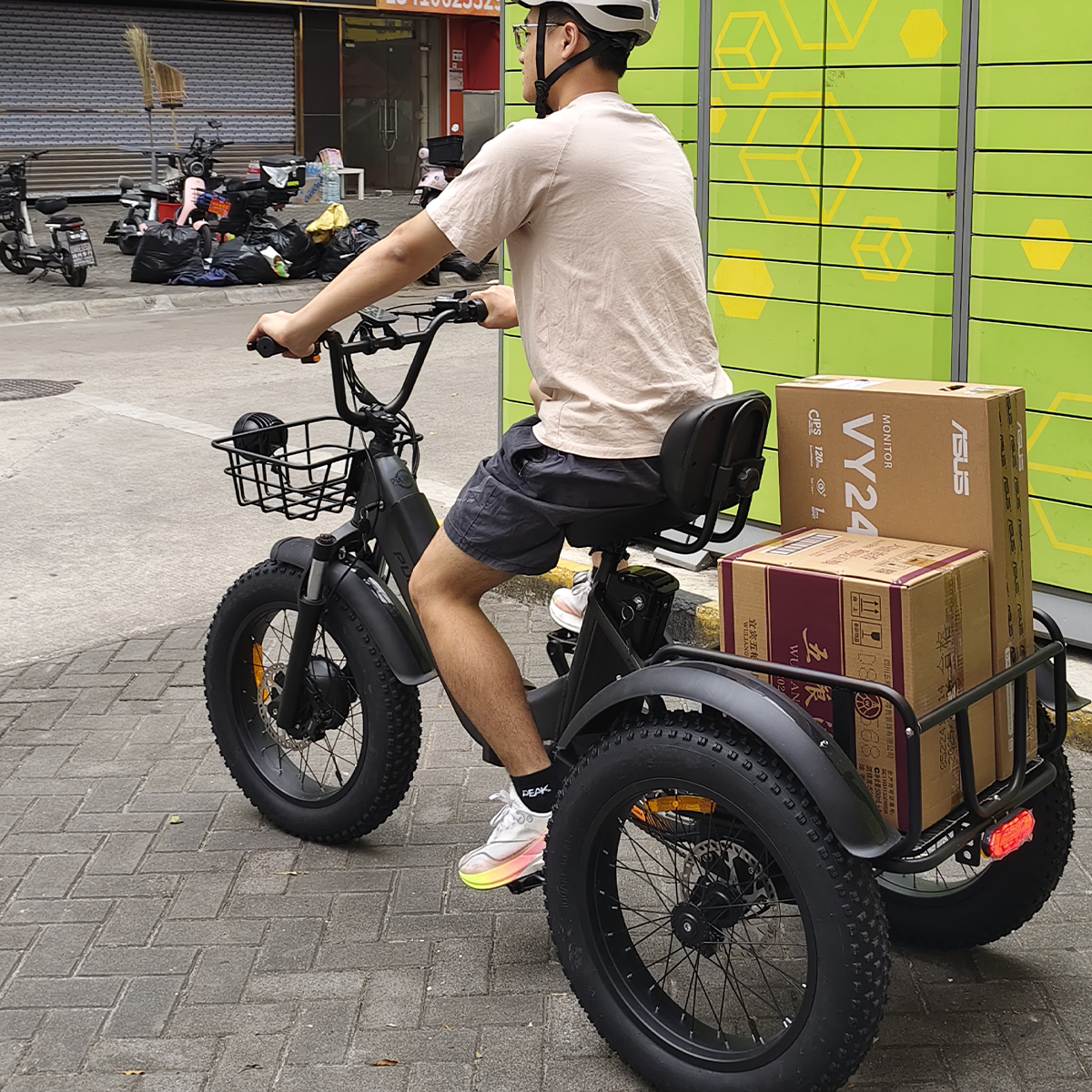 Mężczyzna na czarnym trójkołowcu przewozi pudła. Ma na sobie kask i szorty. Tło z zieloną ścianą i zaparkowanymi skuterami.
