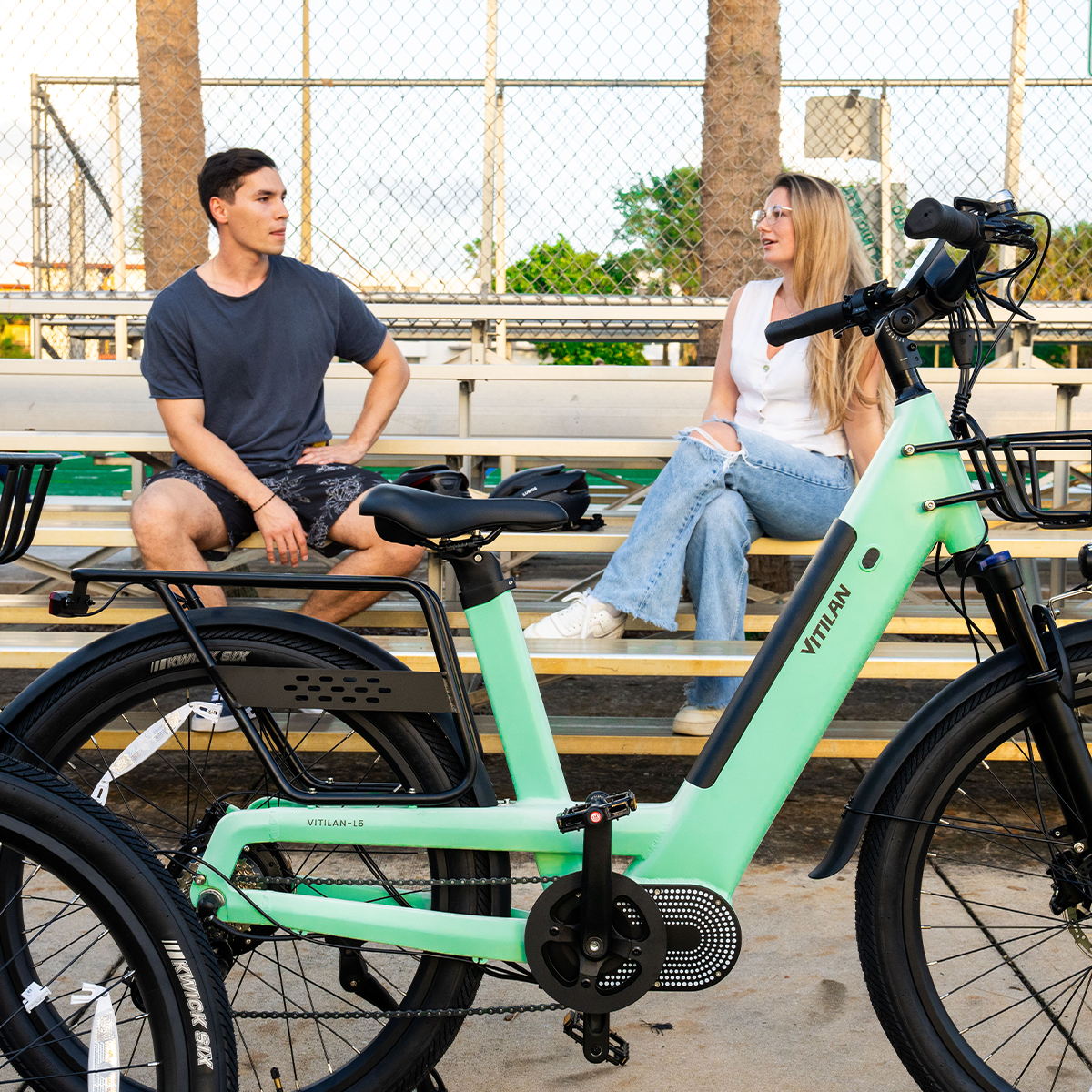 Para siedzi na trybunach obok zielonego roweru elektrycznego. Rower ma czarne opony. W tle palmy.
