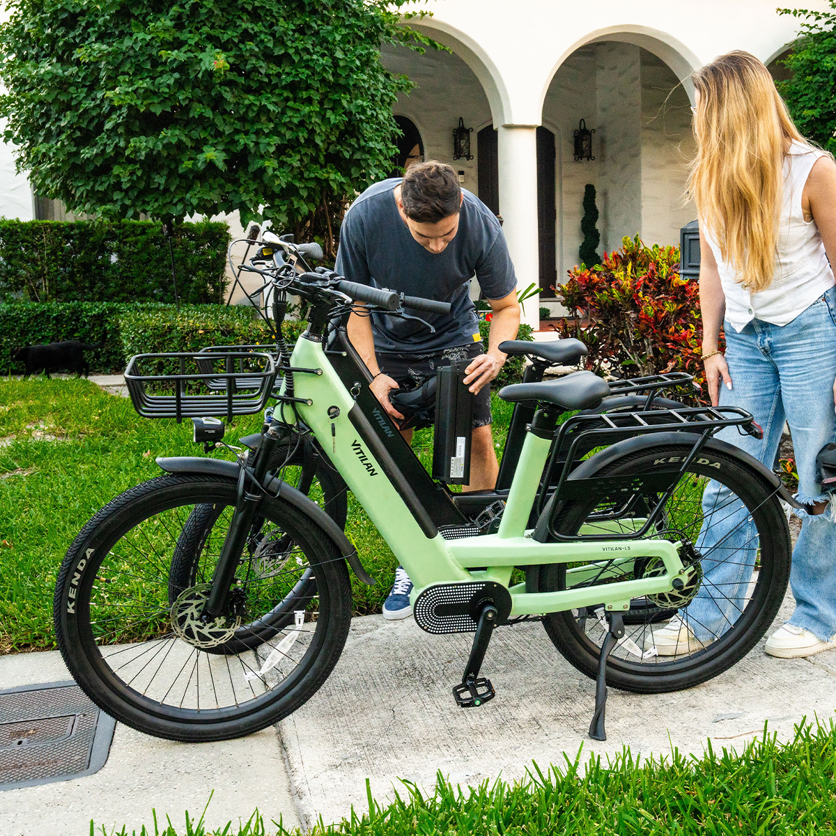 Mężczyzna i kobieta przygotowują rower elektryczny na zewnątrz. Rower jest jasnozielony. Tło stanowi dom i zieleń.