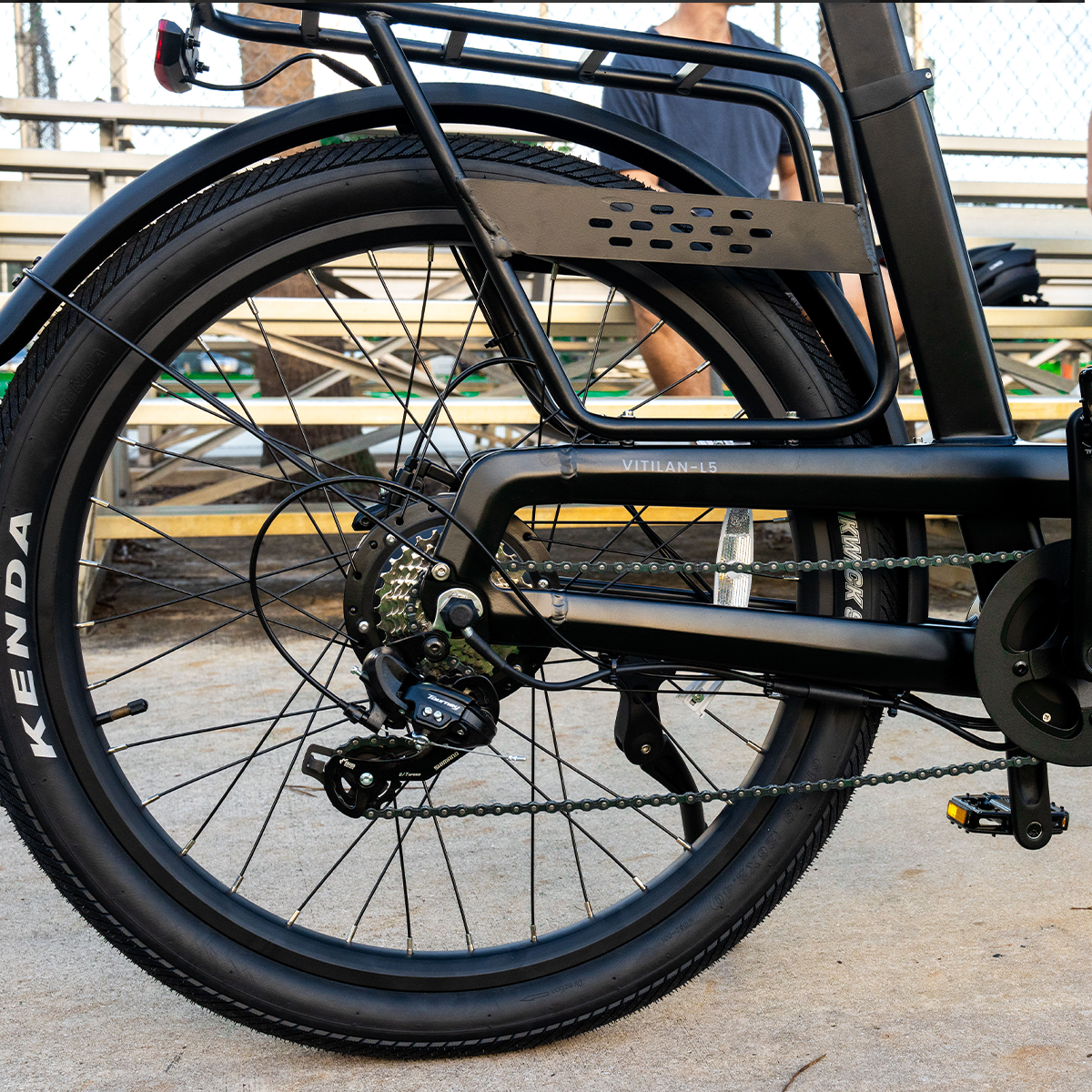 Czarne koło roweru elektrycznego z oponą, łańcuchem, zębatkami i bagażnikiem. Opona Kenda. Osoba w tle.