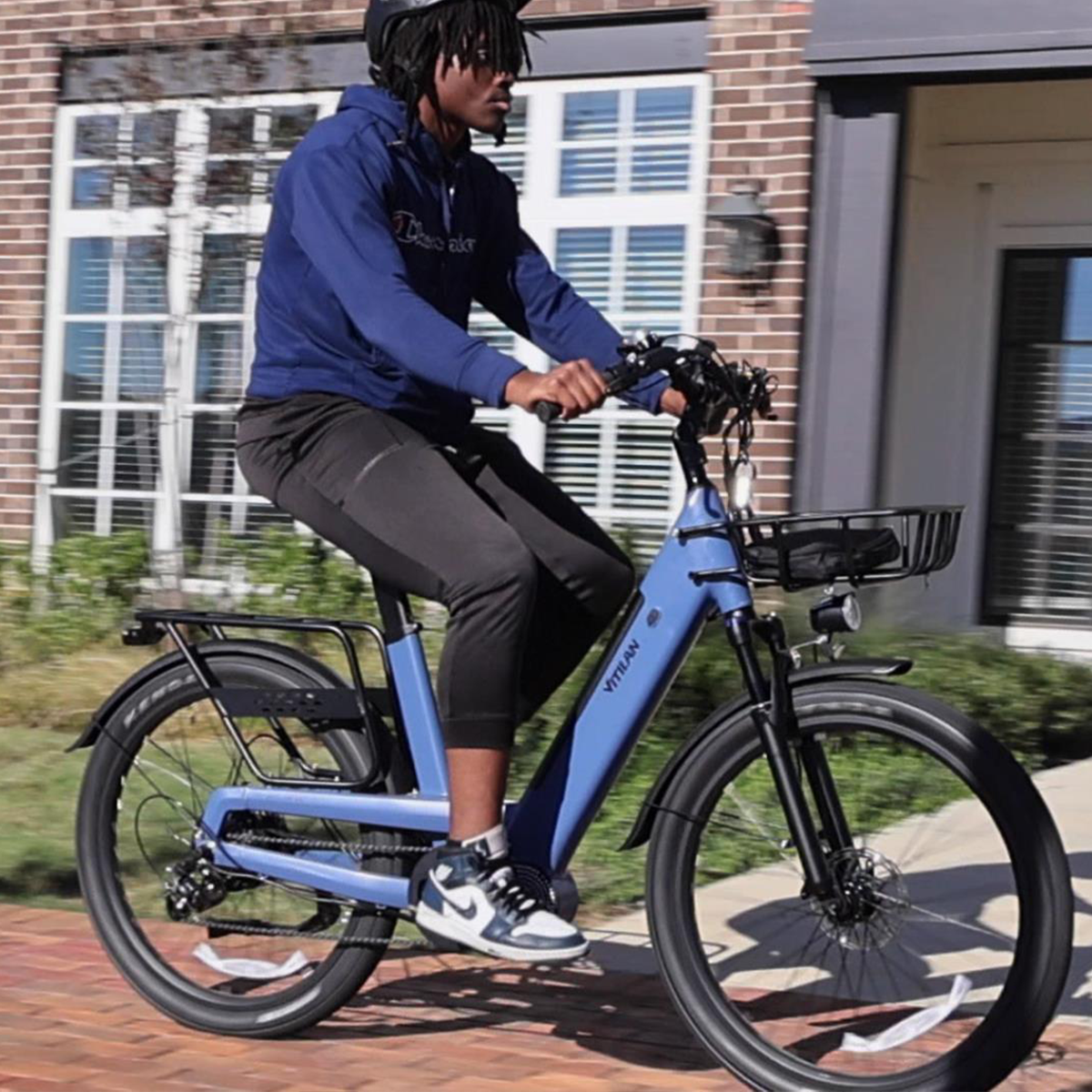 Osoba jedzie niebieskim rowerem elektrycznym. Osoba ma na sobie kask, niebieską bluzę z kapturem, czarne spodnie i białe trampki. Budynek z cegieł w tle.