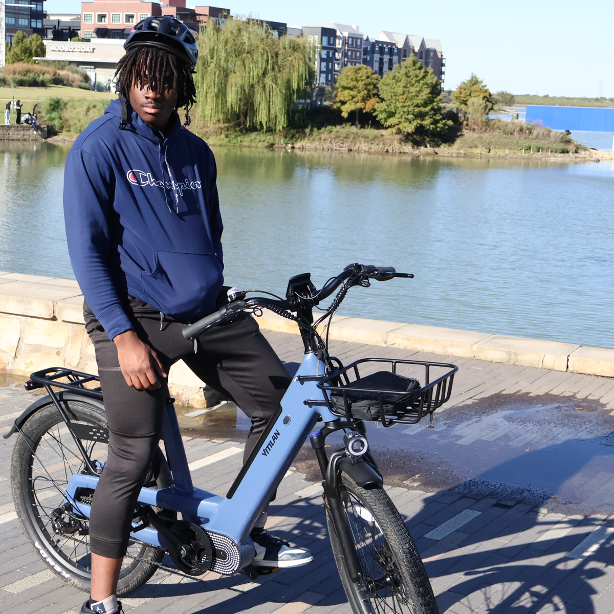 Mężczyzna w niebieskiej bluzie z kapturem Champion i kasku siedzi na niebieskim rowerze Vitilan nad jeziorem. Budynki w tle.