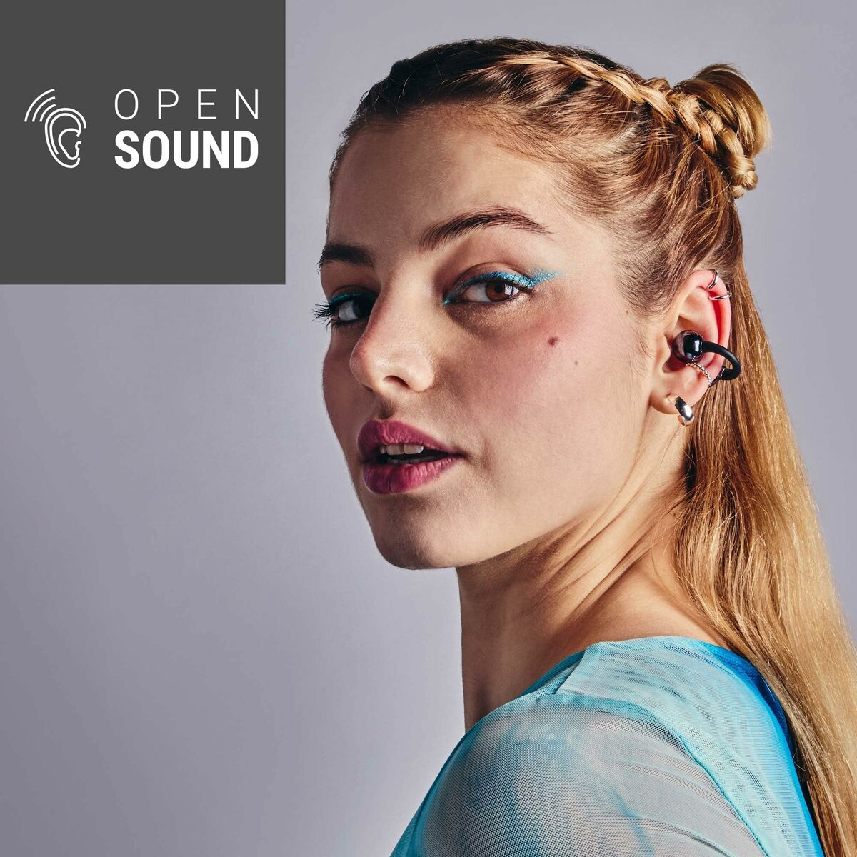 Una donna con i capelli biondi e l'eyeliner blu indossa un apparecchio acustico. L'immagine ha un logo "OPEN SOUND".