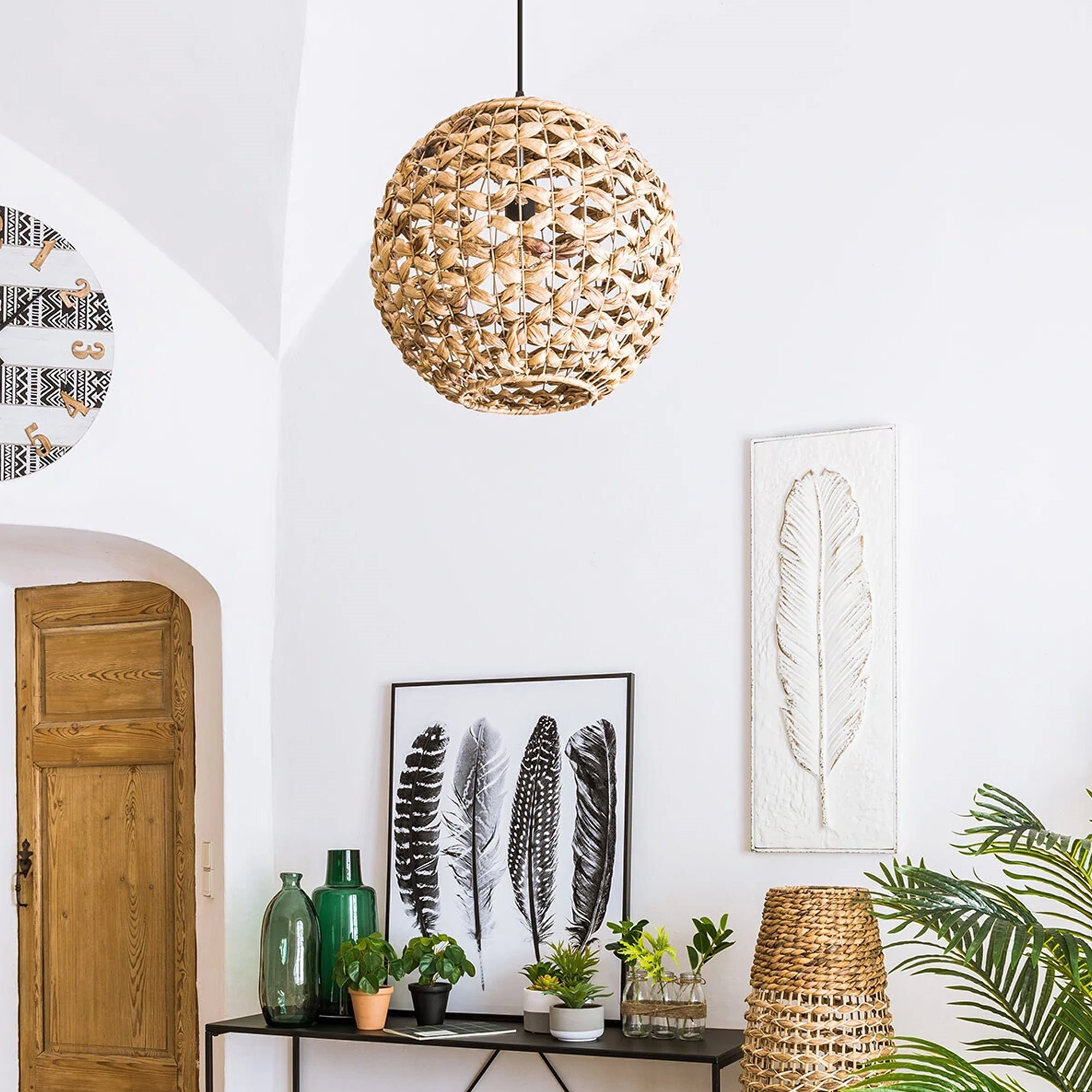 Una habitación con una lámpara colgante tejida, una obra de arte de plumas, una mesa negra con plantas y una puerta de madera.