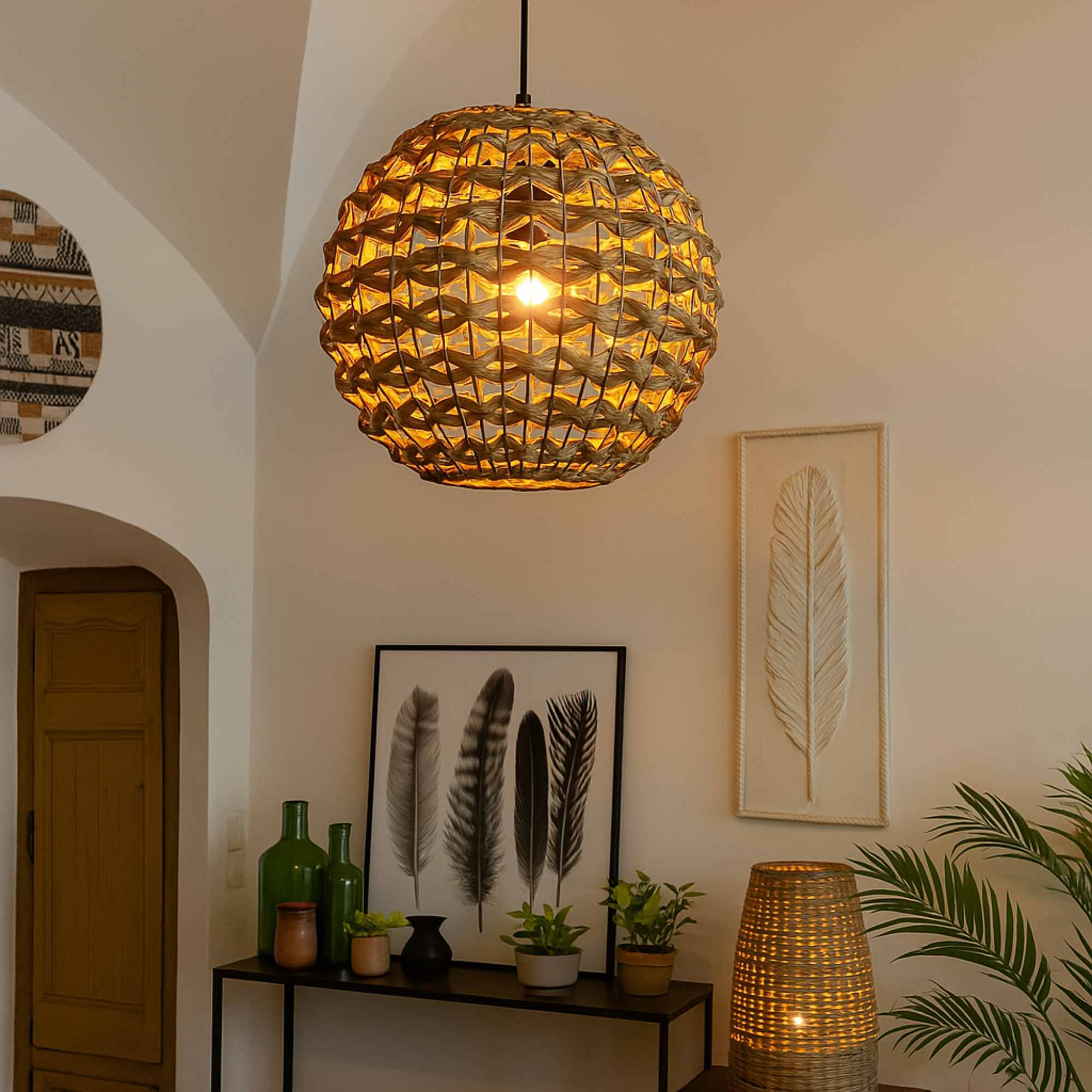 Una lámpara colgante tejida cuelga sobre una mesa con decoración, incluyendo arte de plumas, plantas y una lámpara tejida.