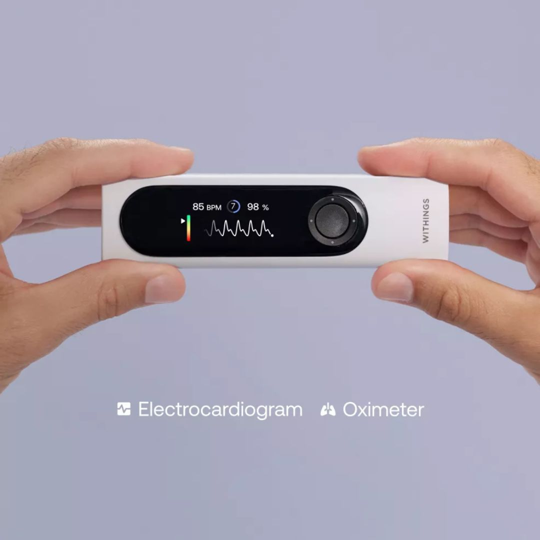 Dłonie trzymają białe urządzenie Withings, wyświetlające tętno (85 BPM) i saturację tlenem (98%). EKG i oksymetr.